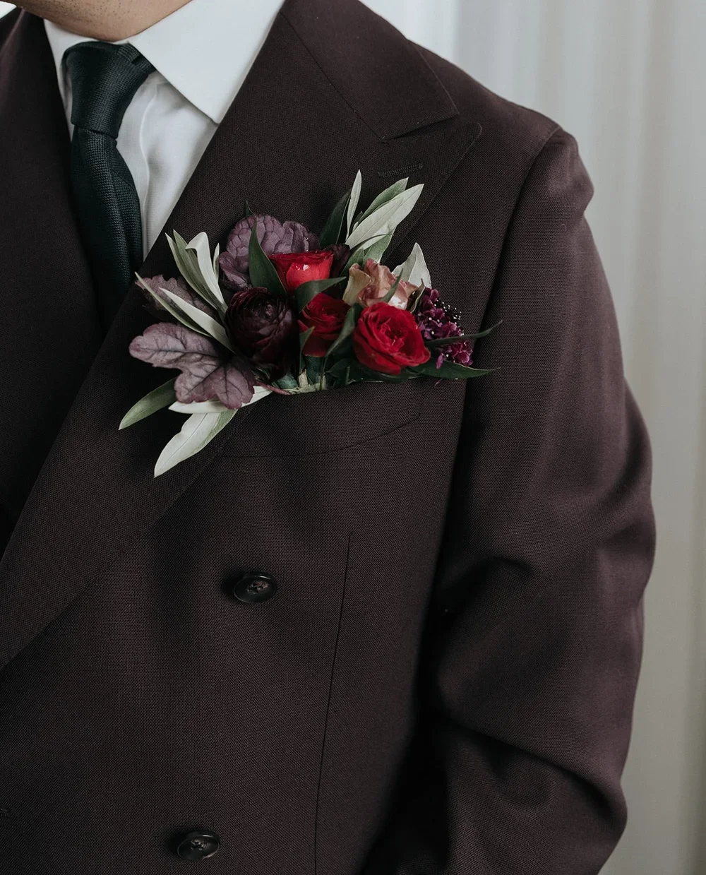 Man wearing a dark suit with a floral boutonniere on his lapel, featuring red, purple, and green flowers and leaves.