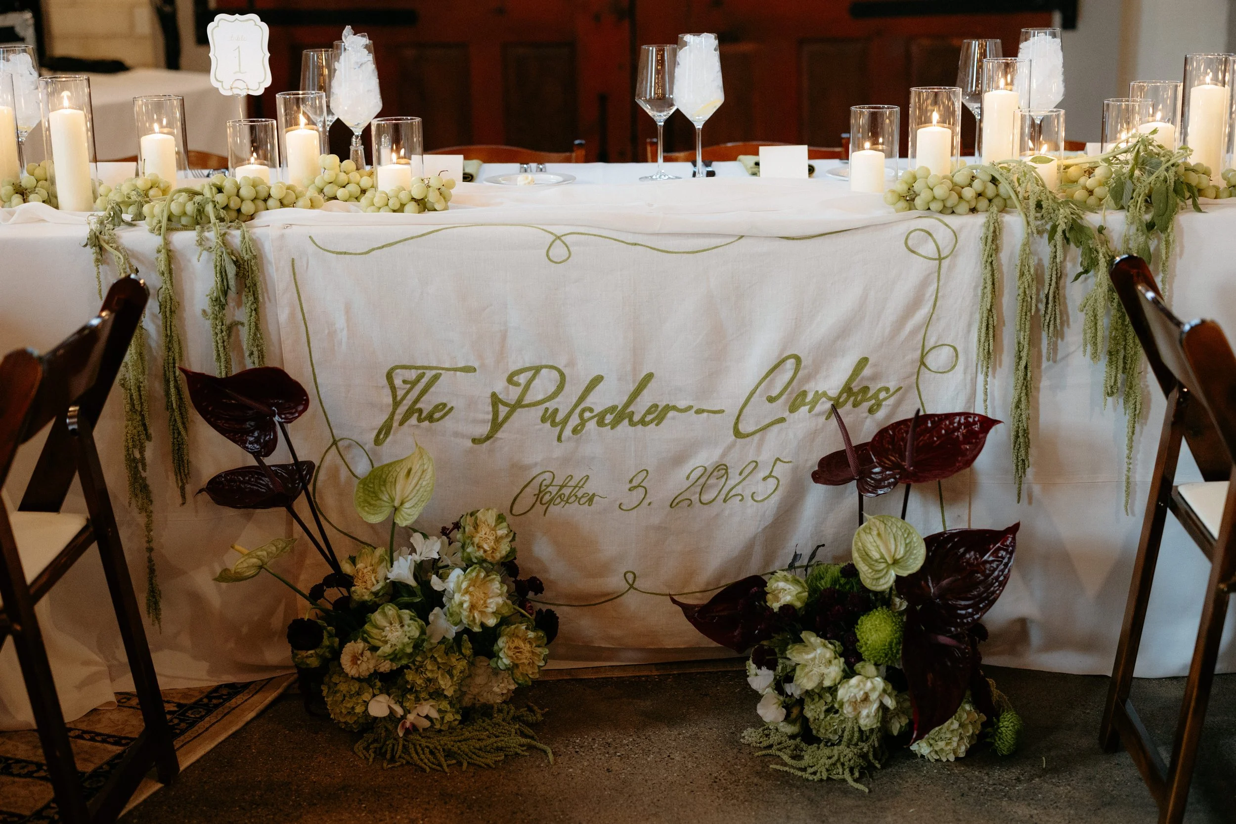 Wedding reception table decorated with white candles in glass holders, white rose and greenery garlands, and floral arrangements at the base. The table has a banner with gold lettering that reads 'The Puscher-Carlos' and the date October 3, 2025. A numbered sign indicating table number 1 is visible. The scene suggests an elegant and festive wedding celebration.