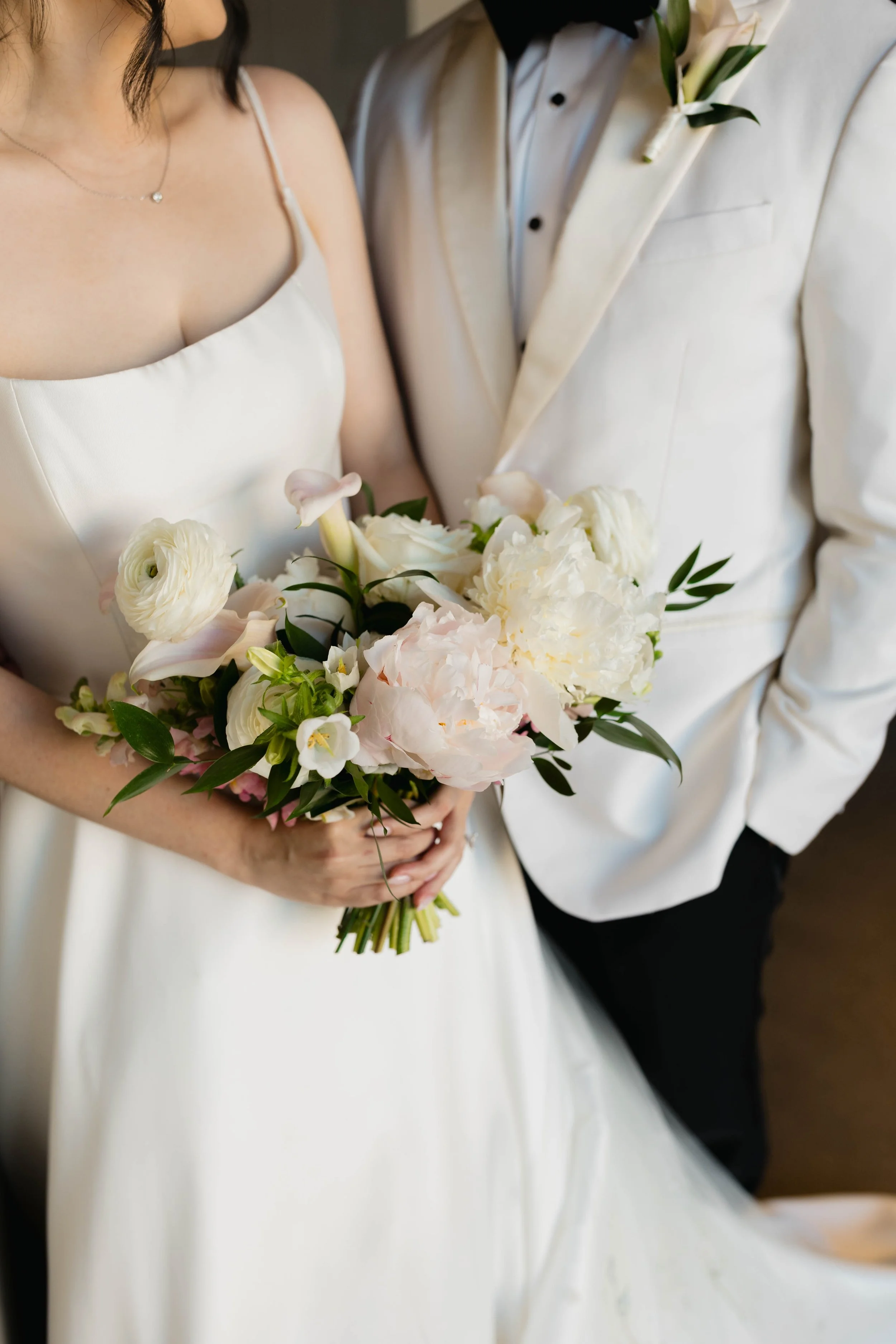 A bride in a white dress holding a bouquet of white and pink flowers, standing next to a groom dressed in a white suit with green leaves on his lapel, at a wedding.