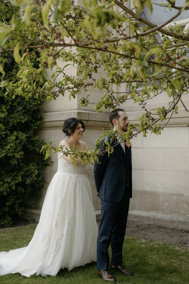 Bride in a white wedding dress playfully pulling a groom's tie outdoors near a stone wall and tree branches.