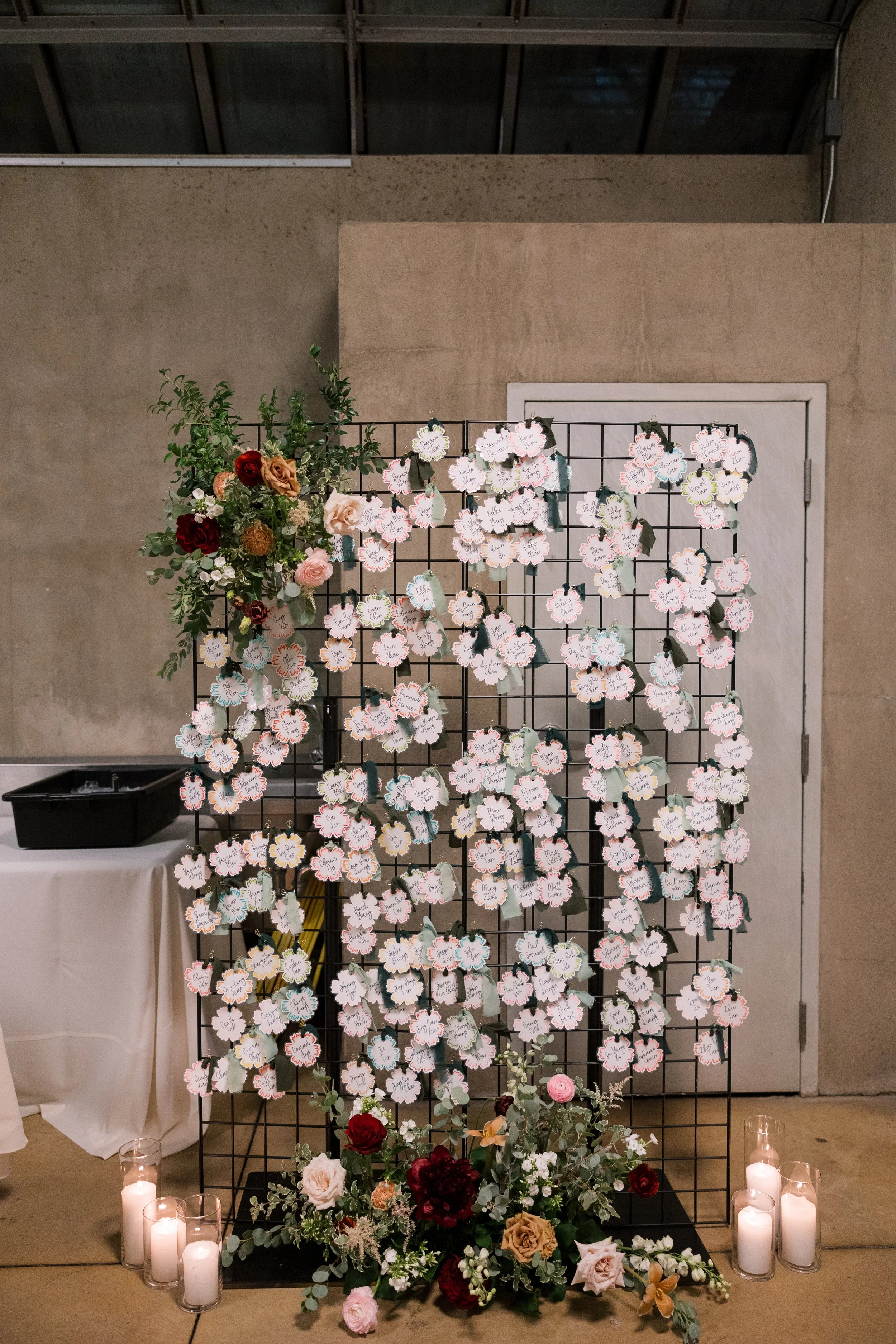 A decorative display featuring a black metal grid with numerous small paper tags attached, each with handwritten names, likely for a wedding or special event. To the left side, there is an arrangement of flowers including roses in shades of red, pink