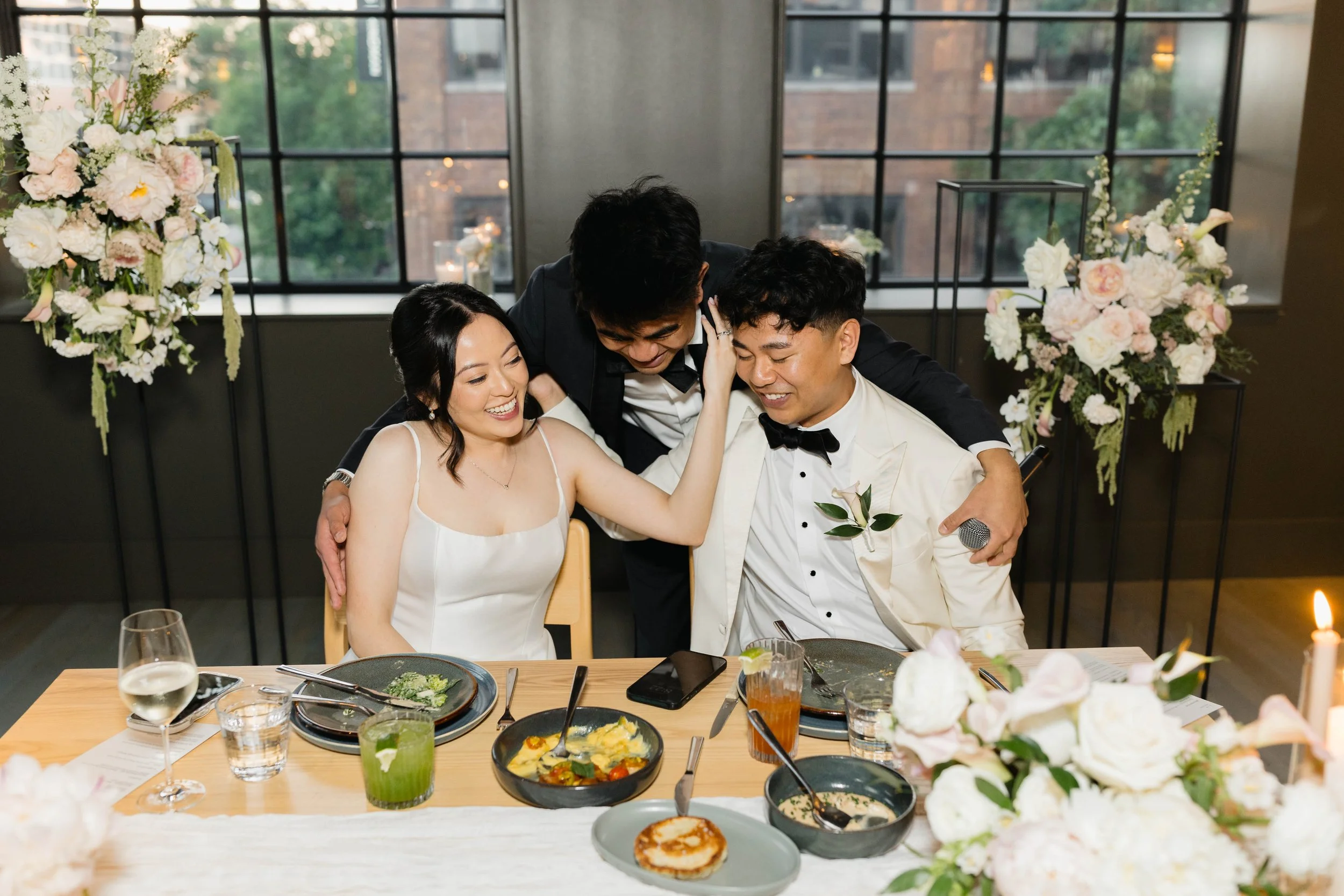 A group of four people, two women and two men, celebrating at a wedding reception, sitting at a table with a floral centerpiece, various dishes, drinks, and a wedding cake, with large windows and city buildings in the background.