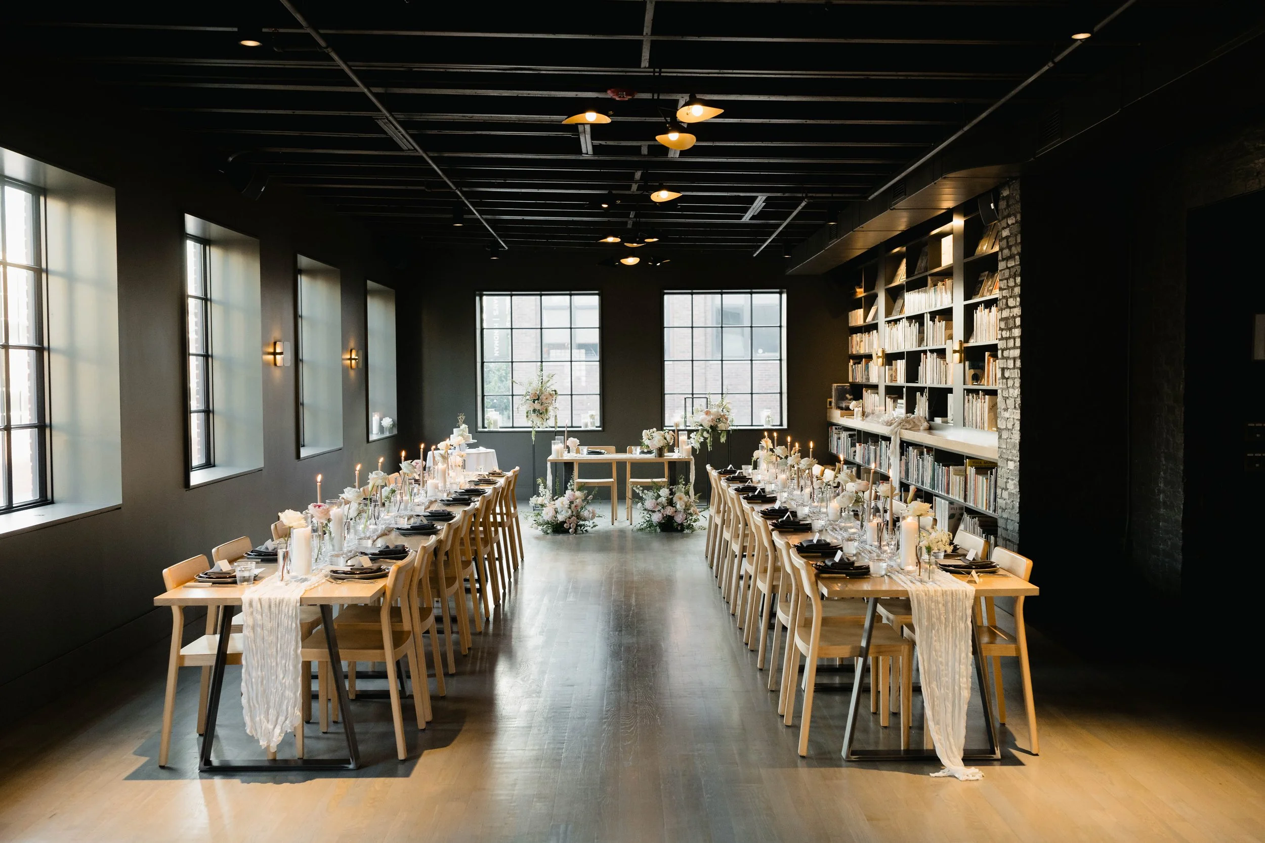 A modern indoor space set up for a wedding reception, featuring long tables with black plates, candles, and floral centerpieces, against a backdrop of large windows and a bookshelf wall.