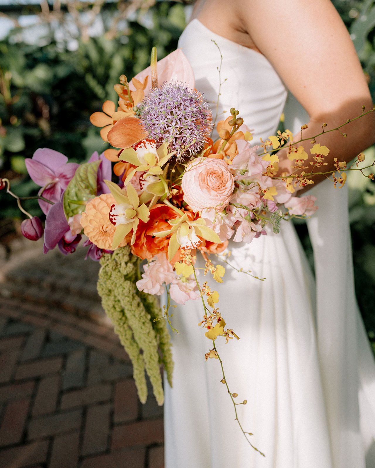 Person in a white dress holding a large colorful bouquet of various flowers, including orchids, roses, and other mixed flowers, in an outdoor setting.