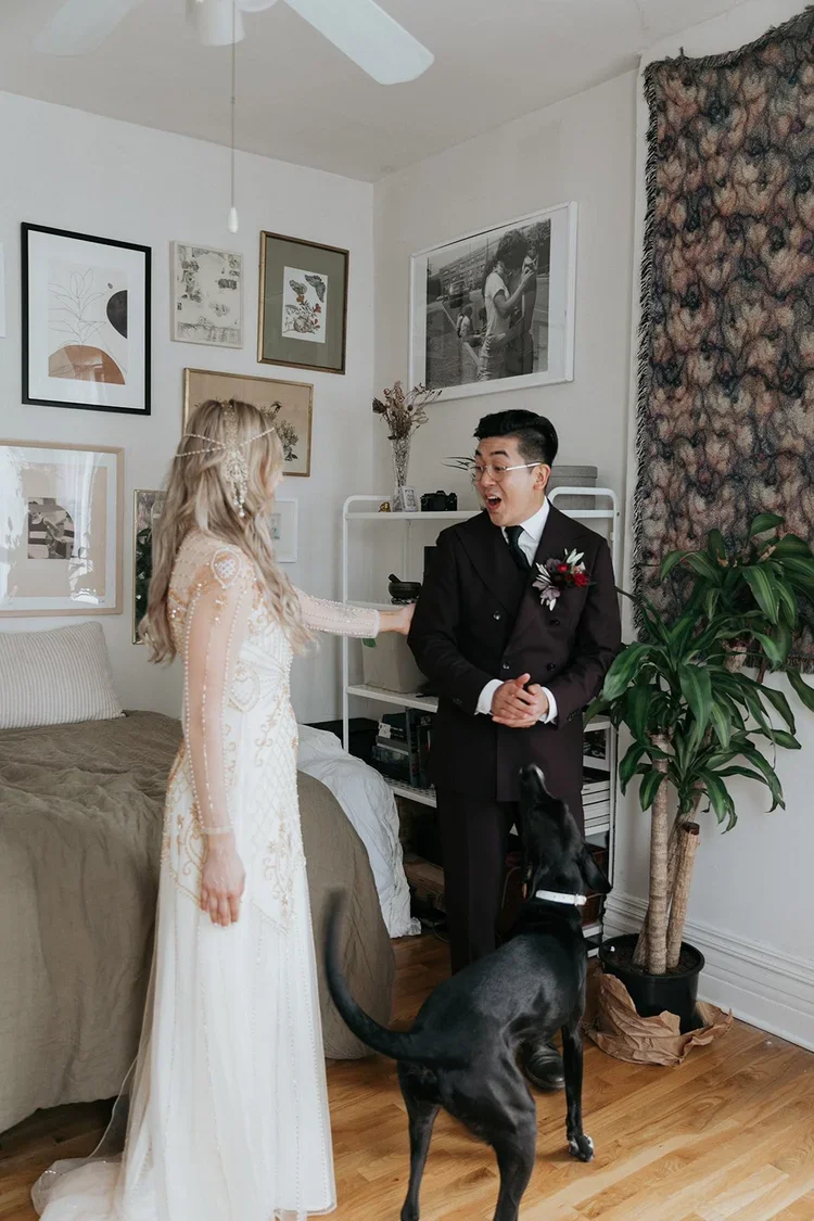 A couple having a wedding moment with the bride in a white wedding dress and the groom in a black suit, interacting with a black dog in a cozy, decorated room.