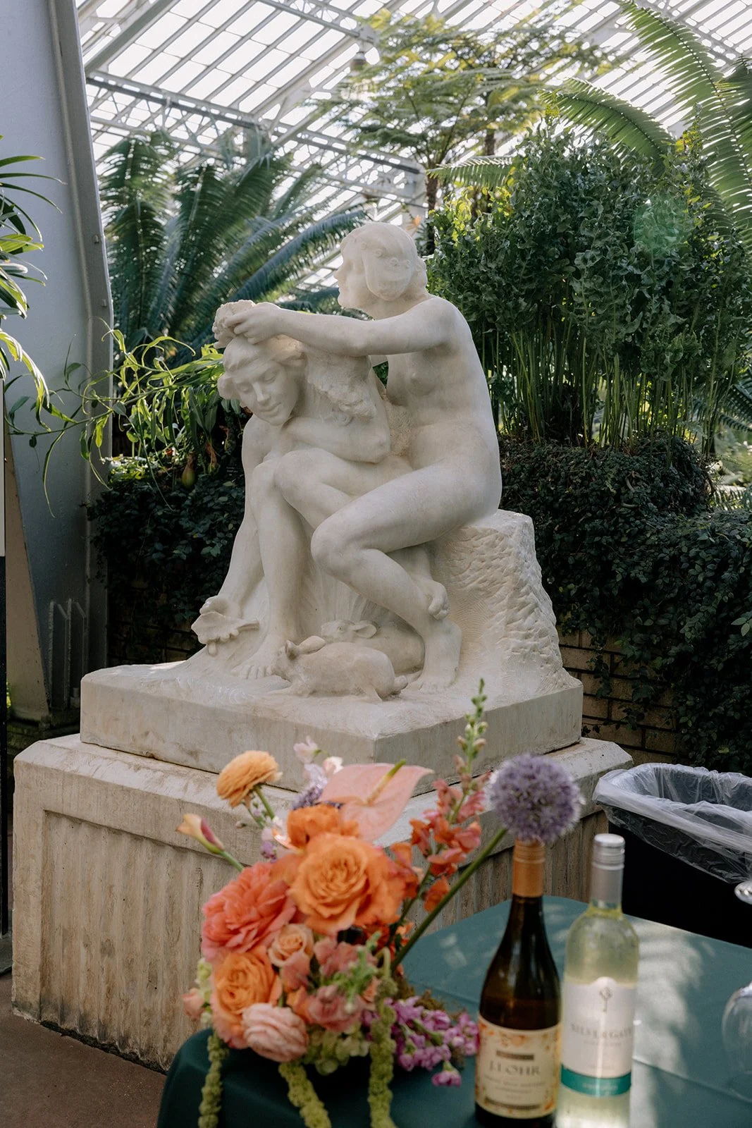 A white marble sculpture of two children, one sitting and one kneeling, with the kneeling child holding the other's head in a garden greenhouse. The sculpture is surrounded by green plants and there are flowers and bottles on a small table in the for
