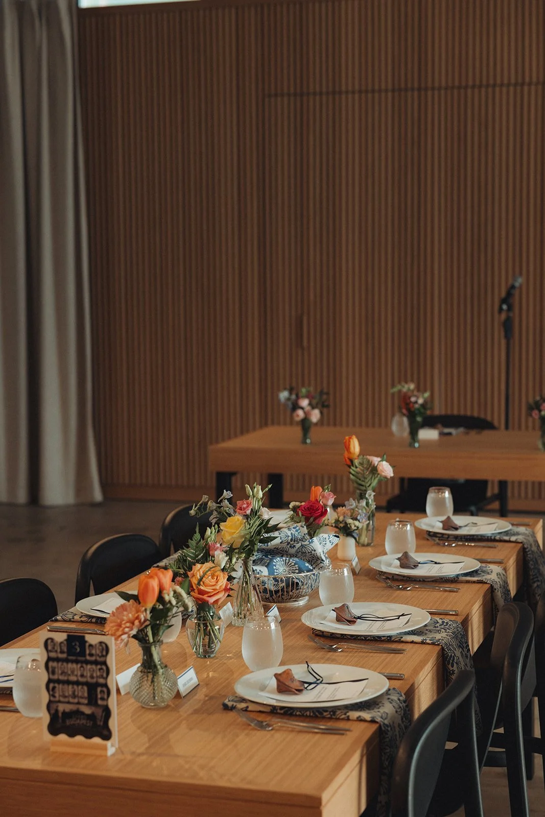 A decorated dining table set for a meal with floral centerpieces, plates, silverware, napkins, and water glasses, in a room with wooden wall paneling and a microphone stand in the background.