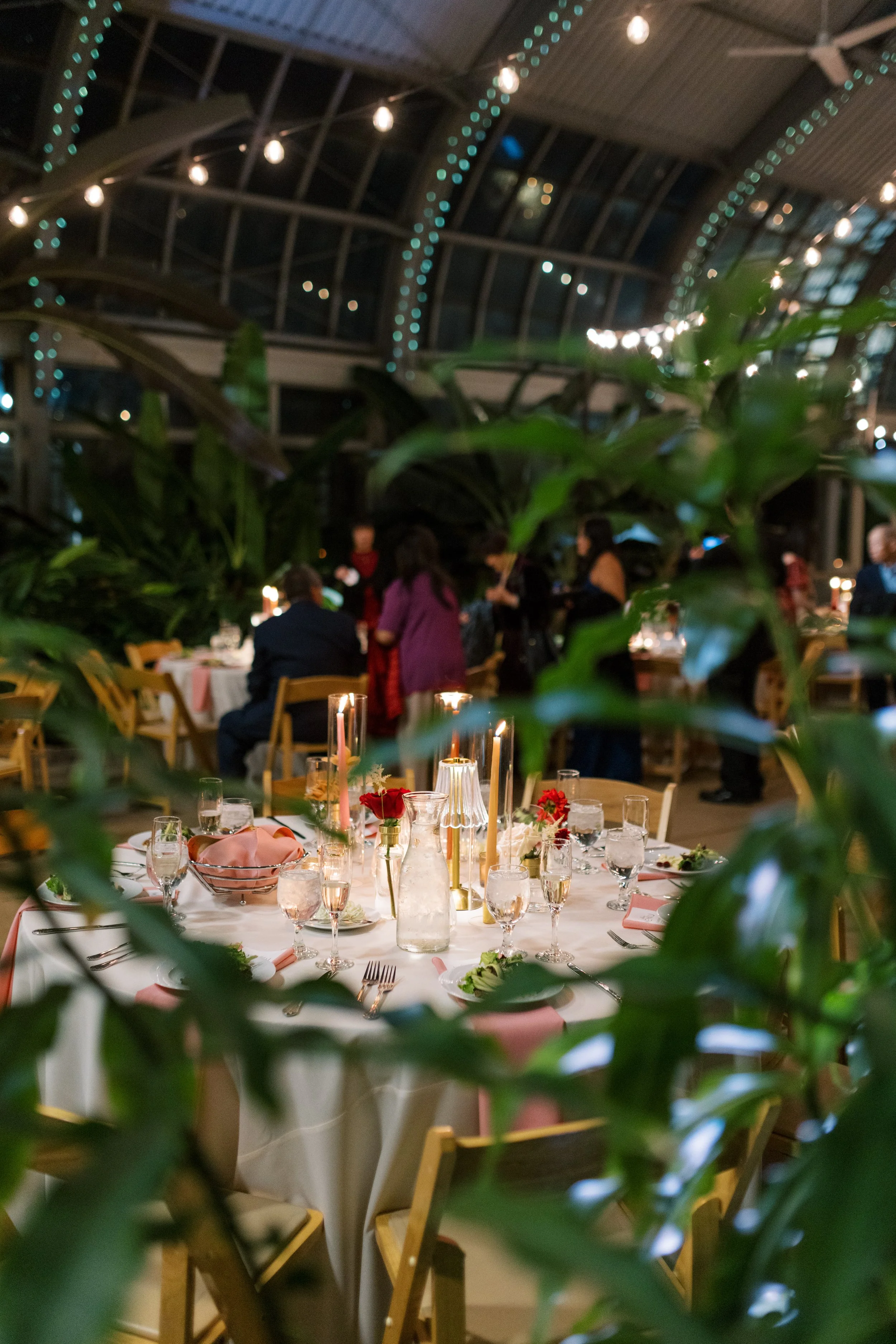 Interior of a banquet hall decorated with string lights, with a round table set for a celebration, featuring candles, flowers, and glassware, partially obscured by green foliage in the foreground.