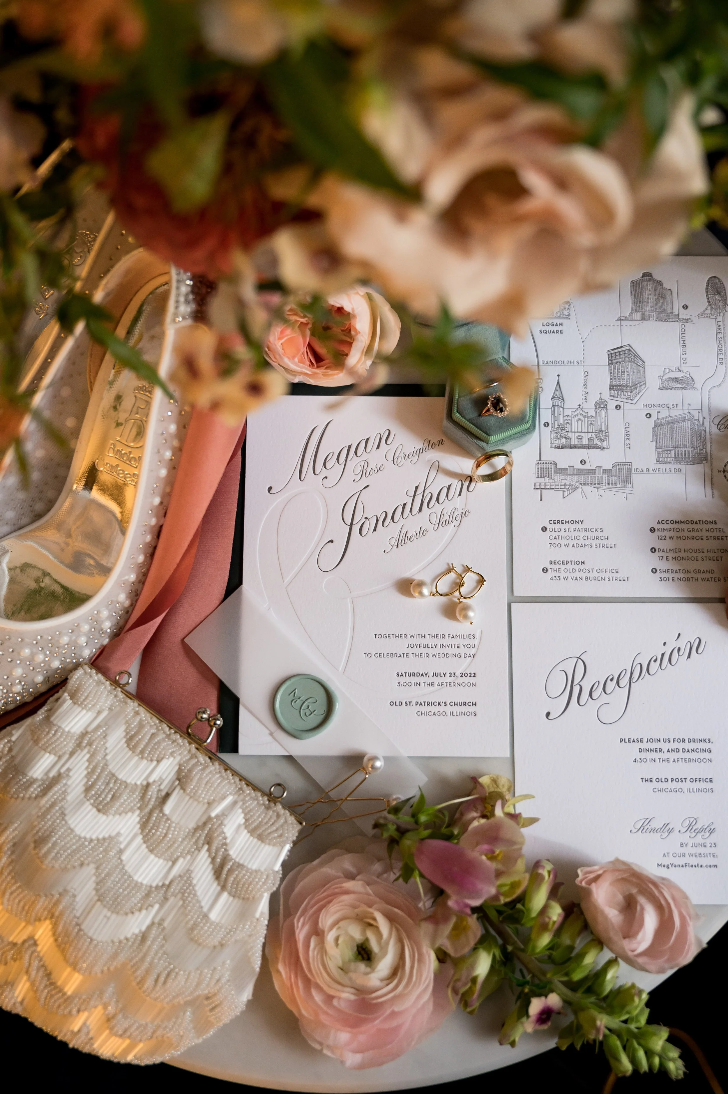 Wedding invitation surrounded by pink and white flowers, a gold ring, pearl jewelry, and decorative items on a white surface.