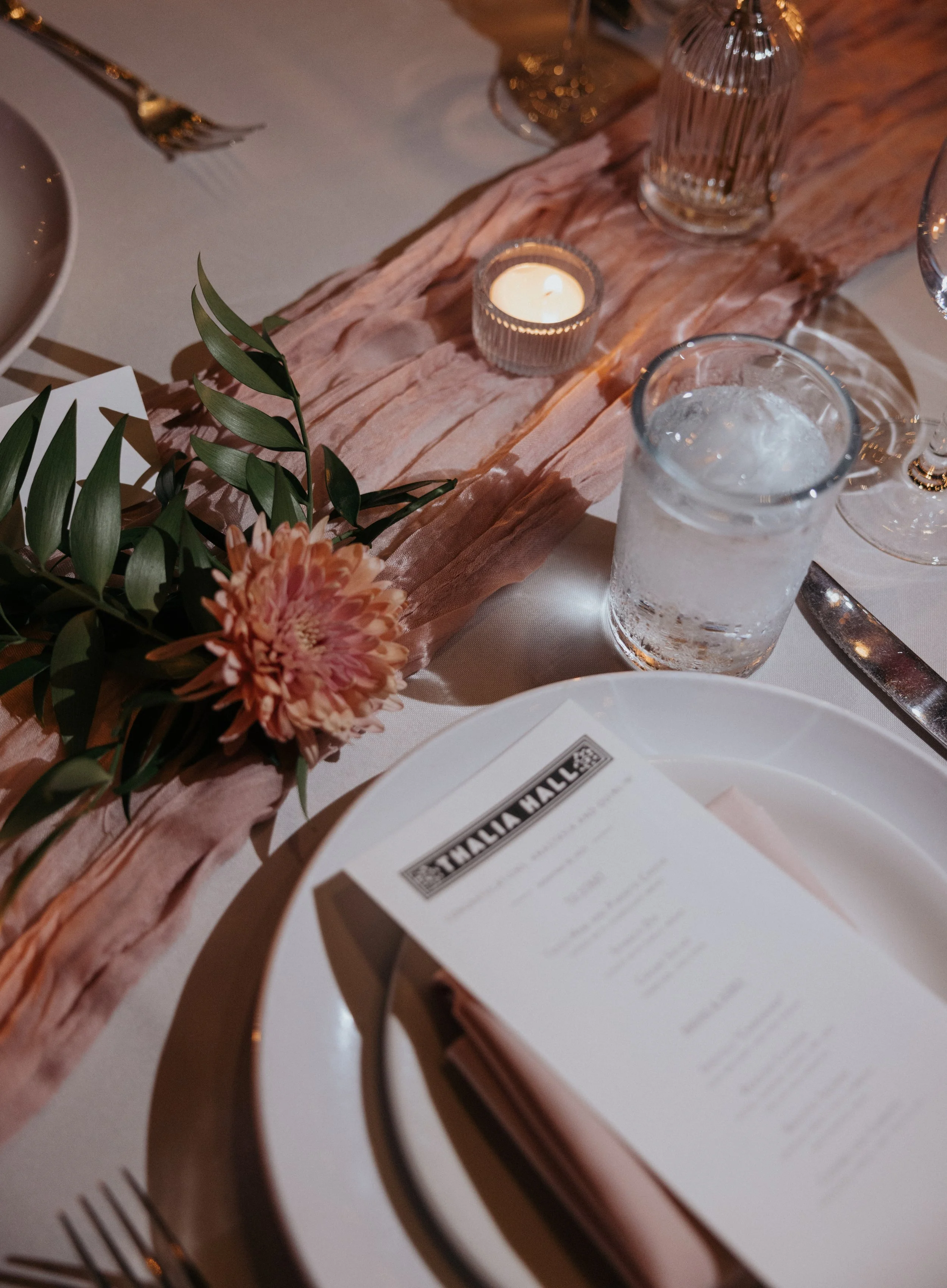 A table setting with a pink floral centerpiece, a small lit candle in a glass holder, a tall ribbed glass vase, a glass of water with ice, and a menu on a white tablecloth. Silverware includes a fork and a knife.