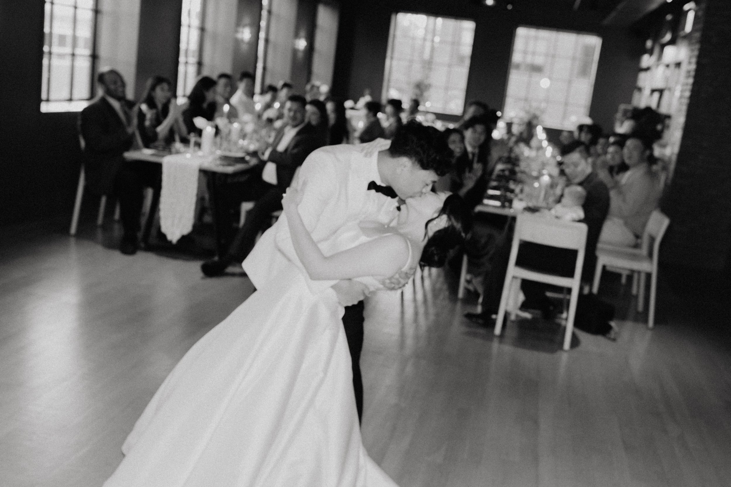 A black-and-white photo of a moment at a wedding reception where a bride and groom are dancing. The bride is wearing a white wedding gown and the groom a dark suit. Guests are seated at tables in the background, watching and celebrating in a decorate