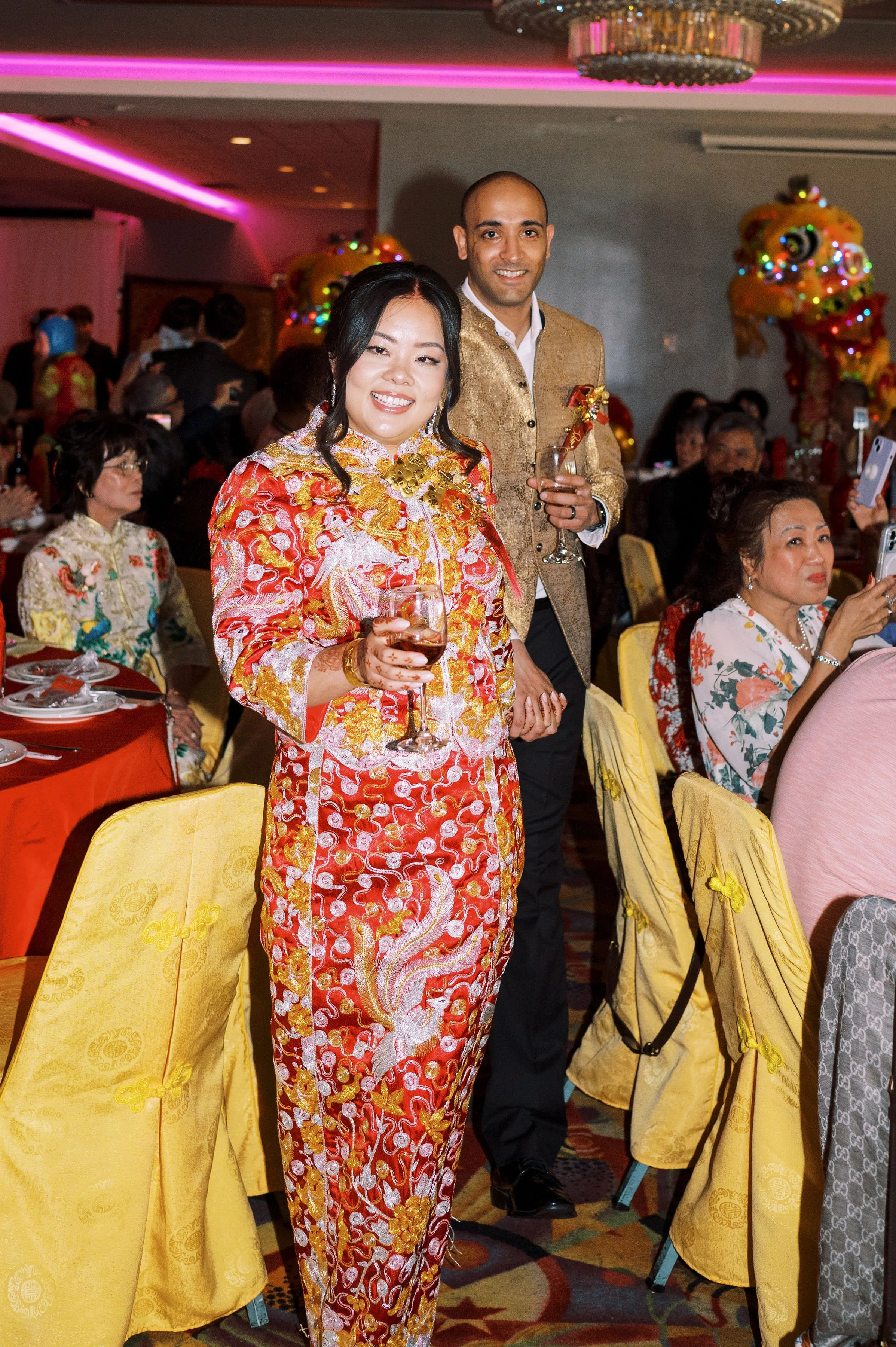 Two people, a woman in a red and gold traditional dress and a man in a gold jacket, are smiling and holding drinks at a festive event with many seated guests and colorful decorations, including a dragon costume in the background.