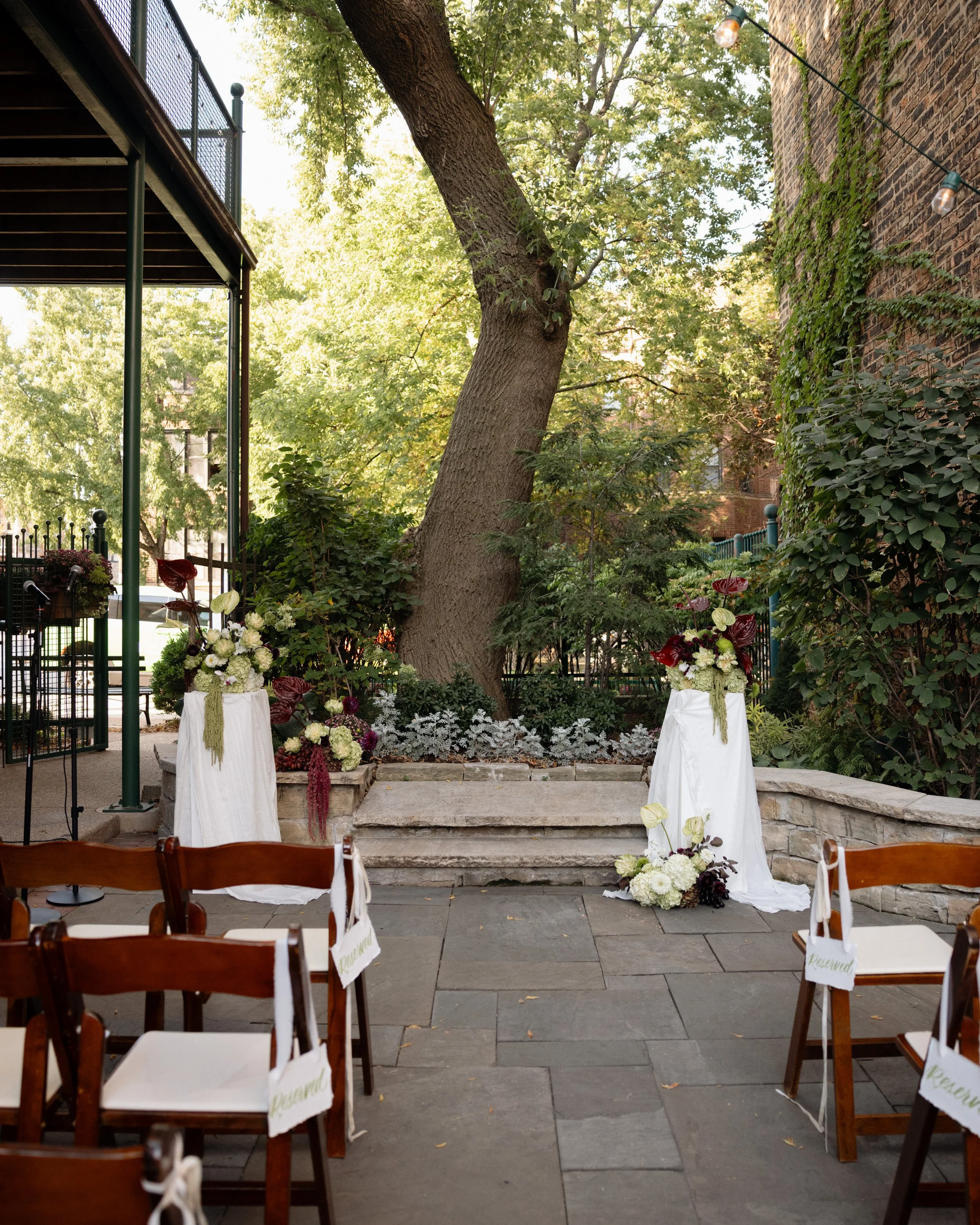 Outdoor wedding ceremony setup with chairs, floral arrangements, and a backdrop of a large tree and brick buildings.