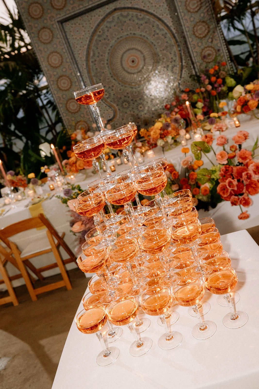 A pyramid of champagne glasses filled with a pink sparkling beverage at a decorated event with floral arrangements and candles.