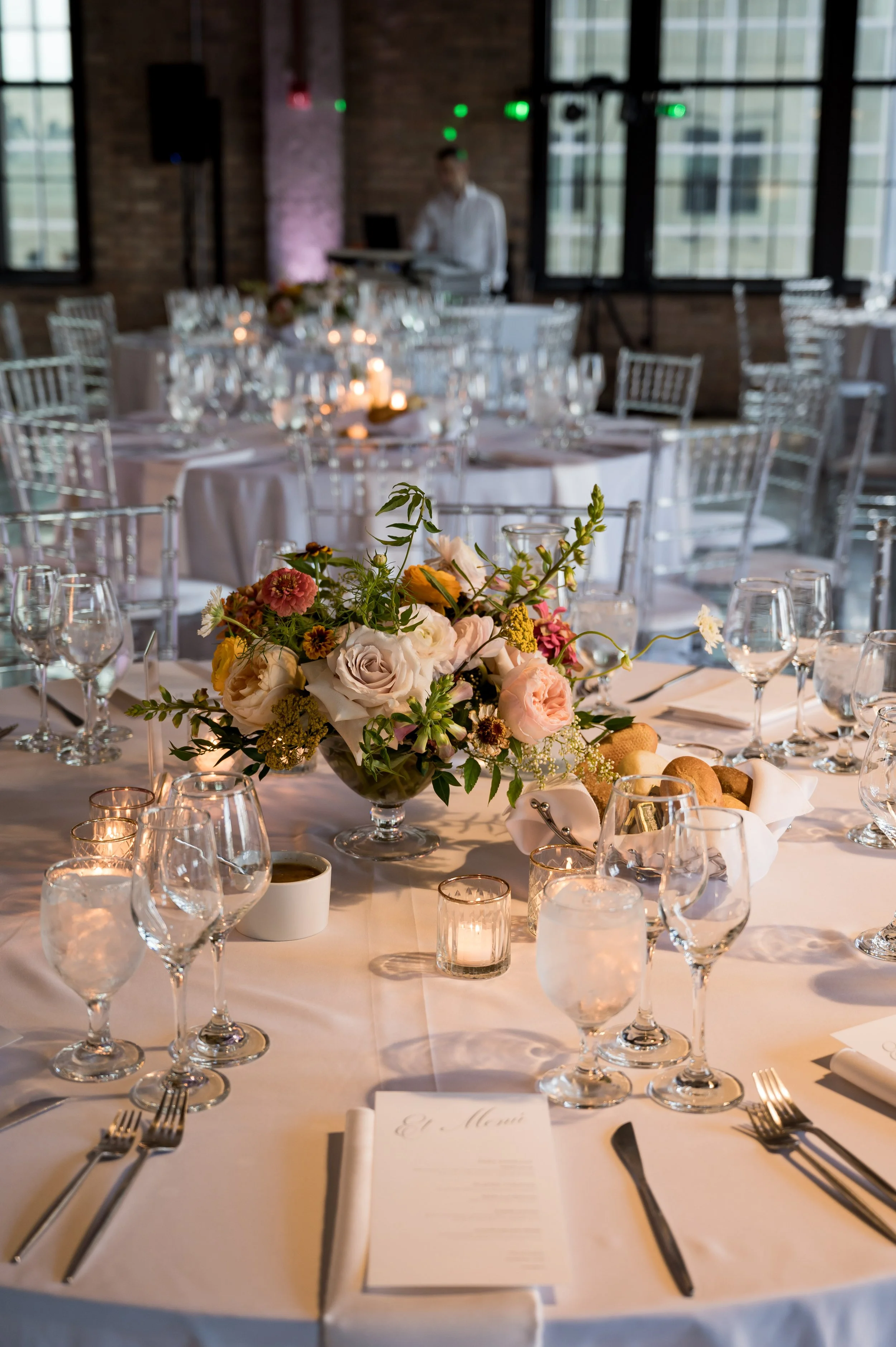 A decorated banquet table at a wedding reception with floral centerpiece, candles, glassware, and a menu, set in a rustic brick-walled venue with large windows. A musician is in the background near a DJ setup.