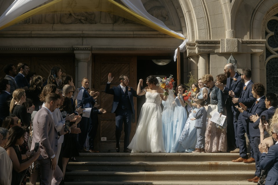 Bride and groom celebrating wedding on church steps, surrounded by guests clapping and cheering, with the bride holding a bouquet of flowers.