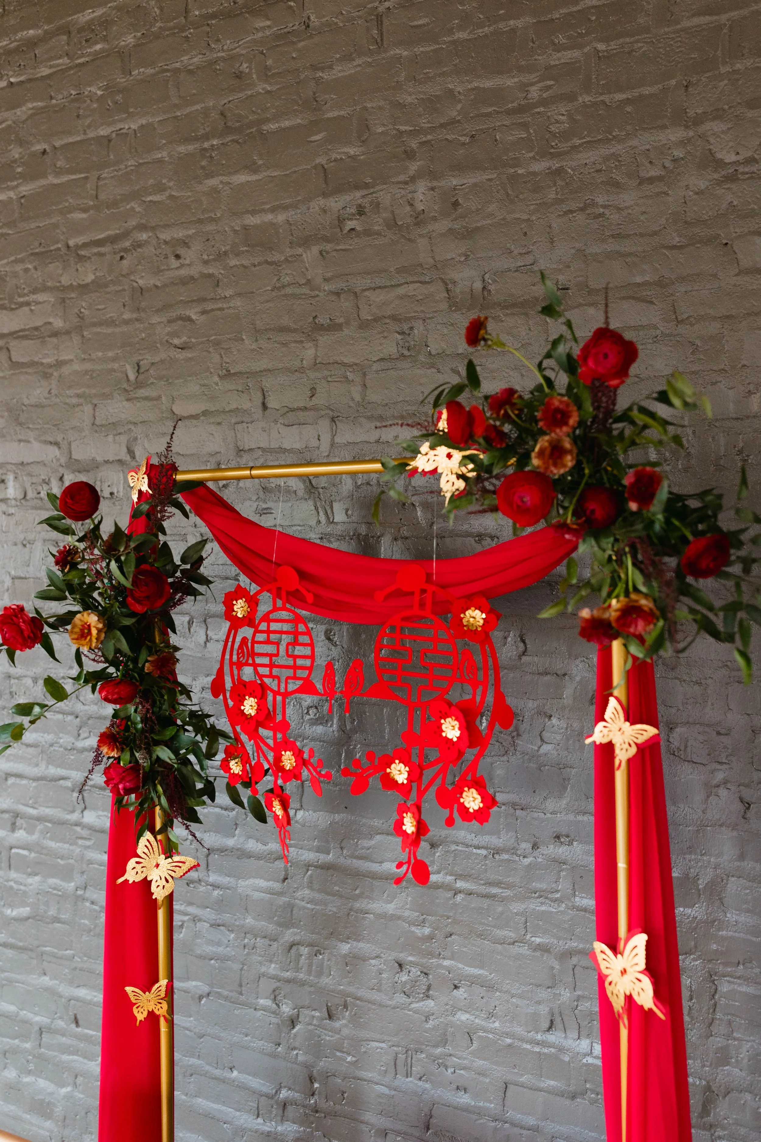 A decorative floral arrangement with red flowers and green leaves, along with gold butterfly ornaments, adorning a red fabric backdrop with hanging paper cutouts of traditional Chinese lanterns and butterflies, set against a gray brick wall.