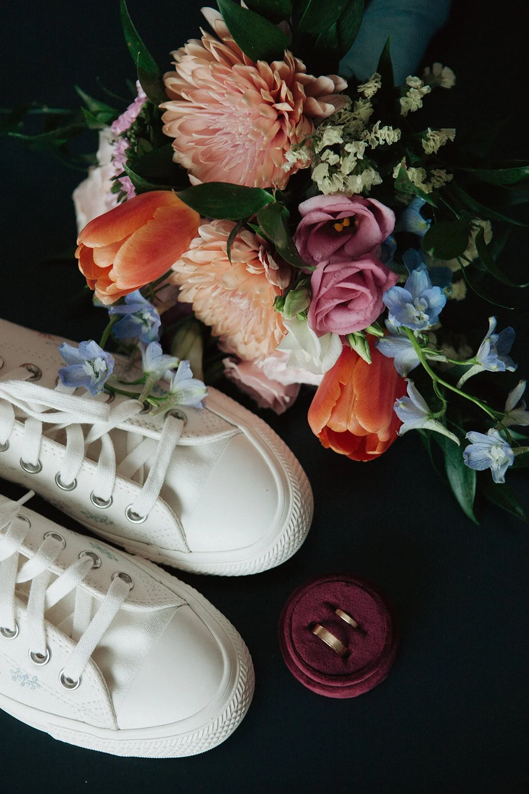 White sneakers with white laces, a red velvet ring box with two gold rings, and a colorful bouquet of flowers including pink roses, orange tulips, light pink dahlias, and blue delphiniums on a black surface.