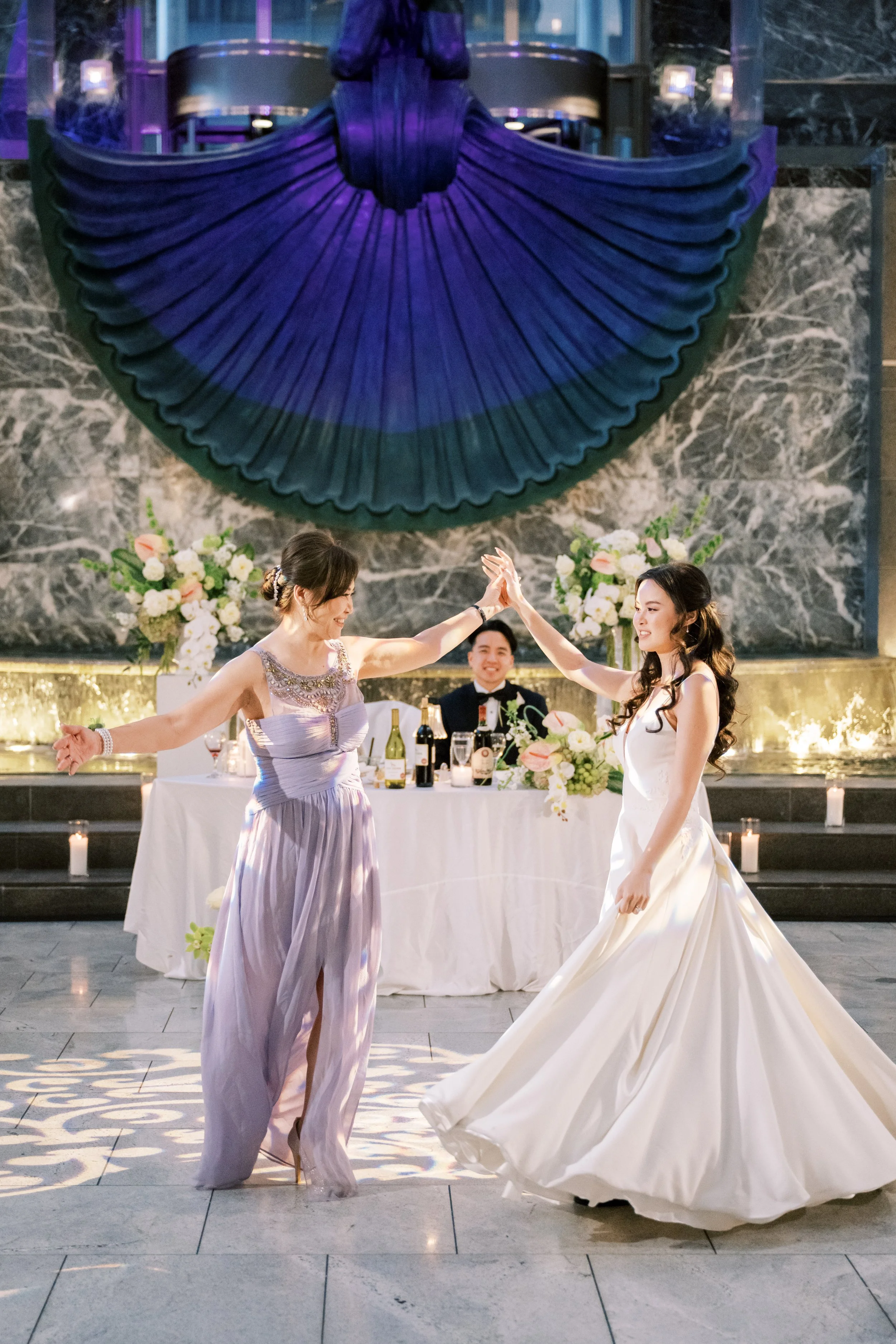Two women dancing at a wedding reception with one wearing a purple dress and the other in a white wedding gown. There are floral arrangements and a marble wall with a large decorative blue and purple art piece in the background.