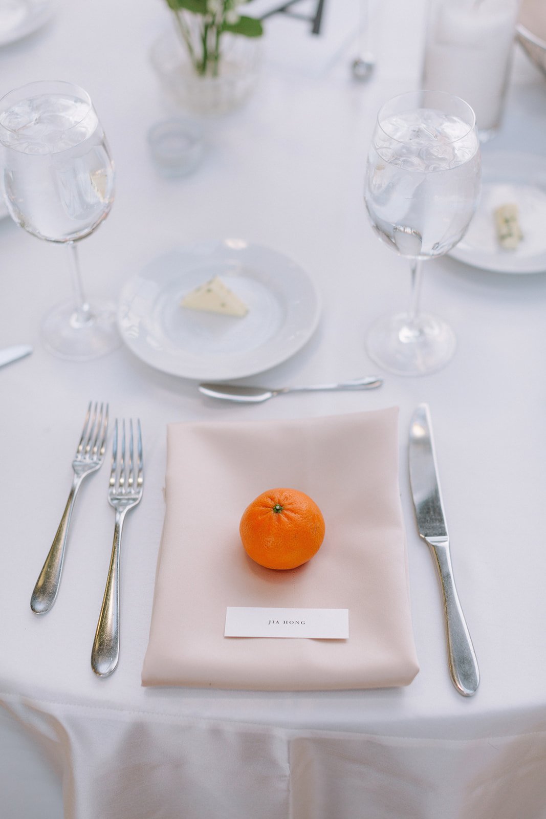 A place setting on a white table with a beige napkin and a mandarin orange on a name card that reads 'Jia Hong'. The setting includes silverware, two glasses of water, and plates with cheese.