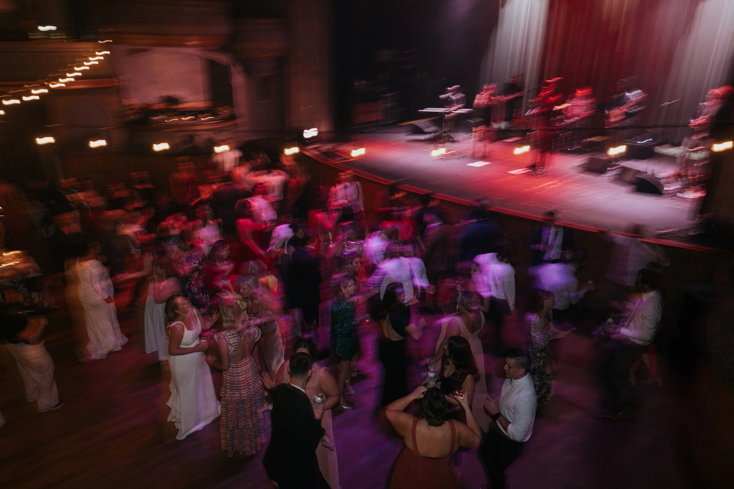 A crowded dance floor with people dancing and socializing, a band performing on stage with musicians playing guitars and keyboards, stage lights creating colorful effects, and a rustic interior with wooden floors and walls.