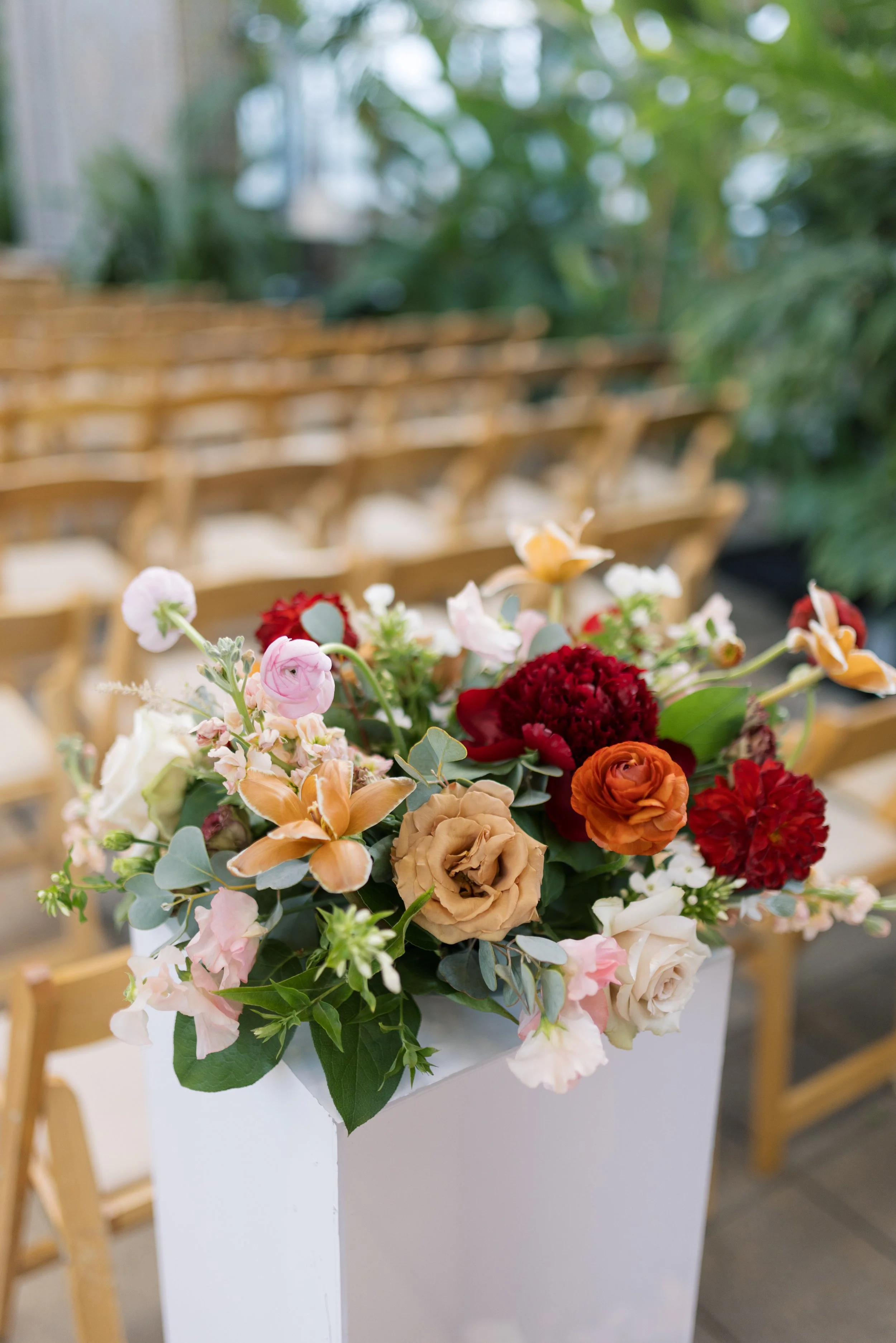 A flower arrangement with pink, red, orange, and cream flowers and green foliage in a white rectangular vase placed on a table, with wooden chairs and greenery in the background.