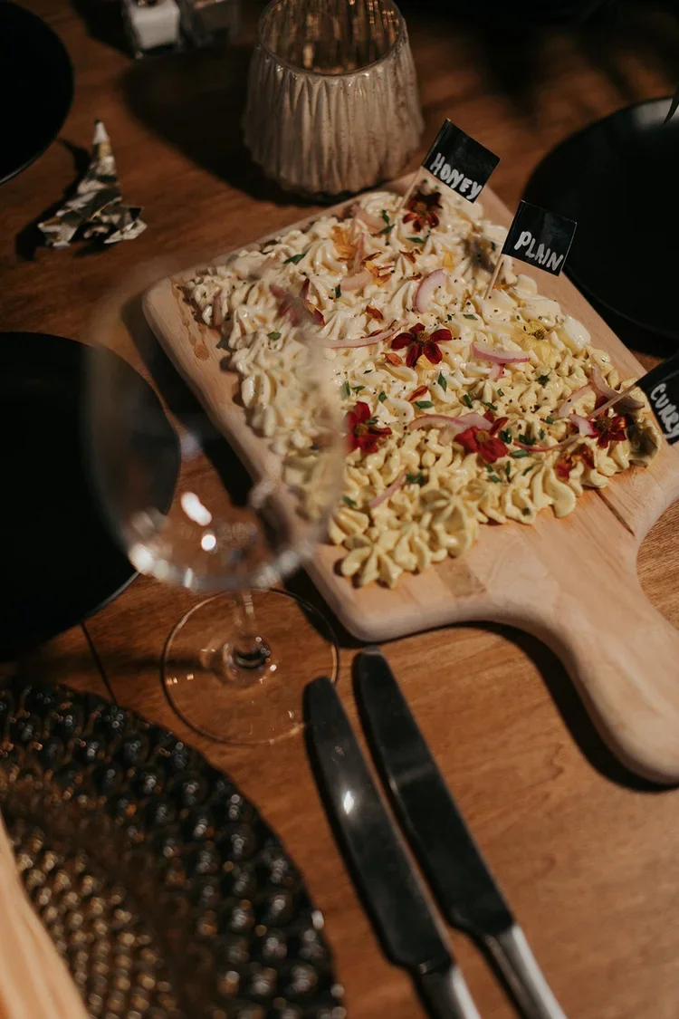 A wooden serving board with an assortment of pasta salad topped with herbs and flowers, on a table with a glass of red wine, a candle holder, and black plates and utensils.