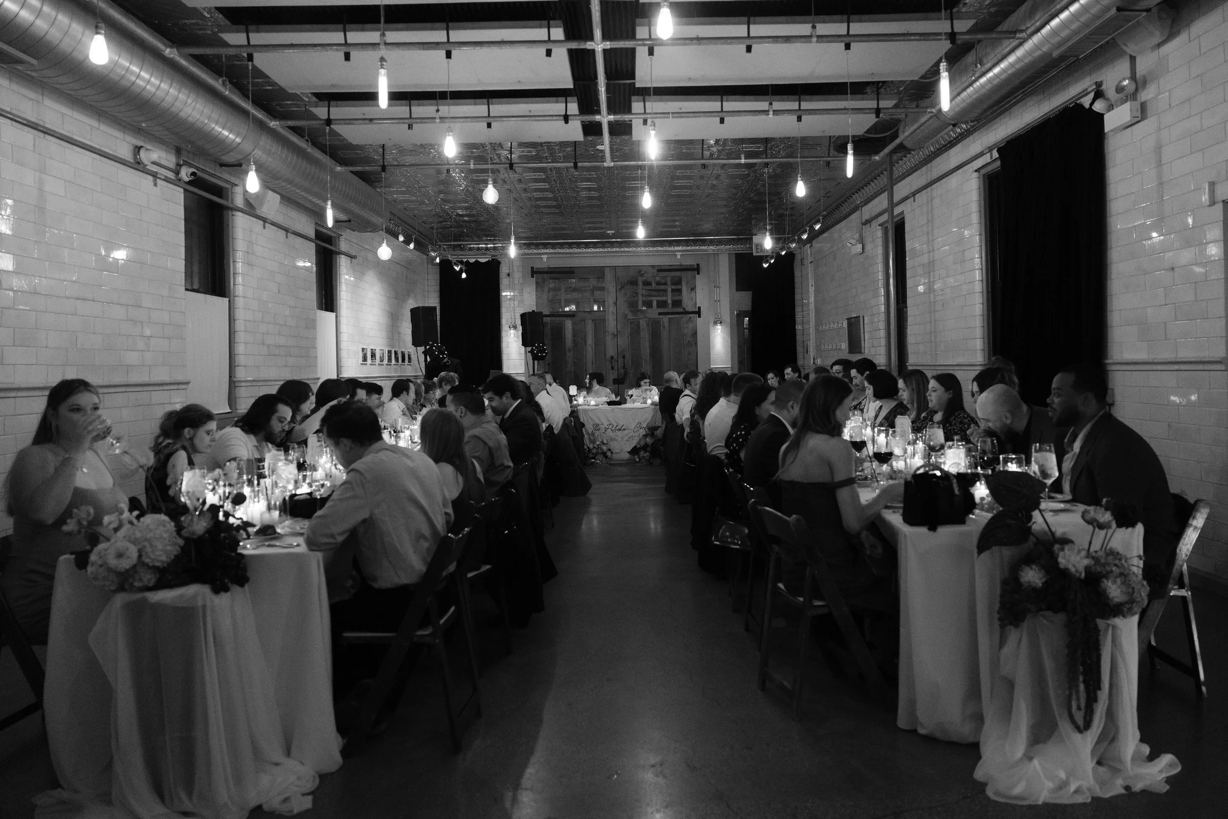A black and white photo of a long banquet hall filled with people seated at decorated tables, lit by hanging exposed bulbs, with brick walls, large windows, and dark curtains.