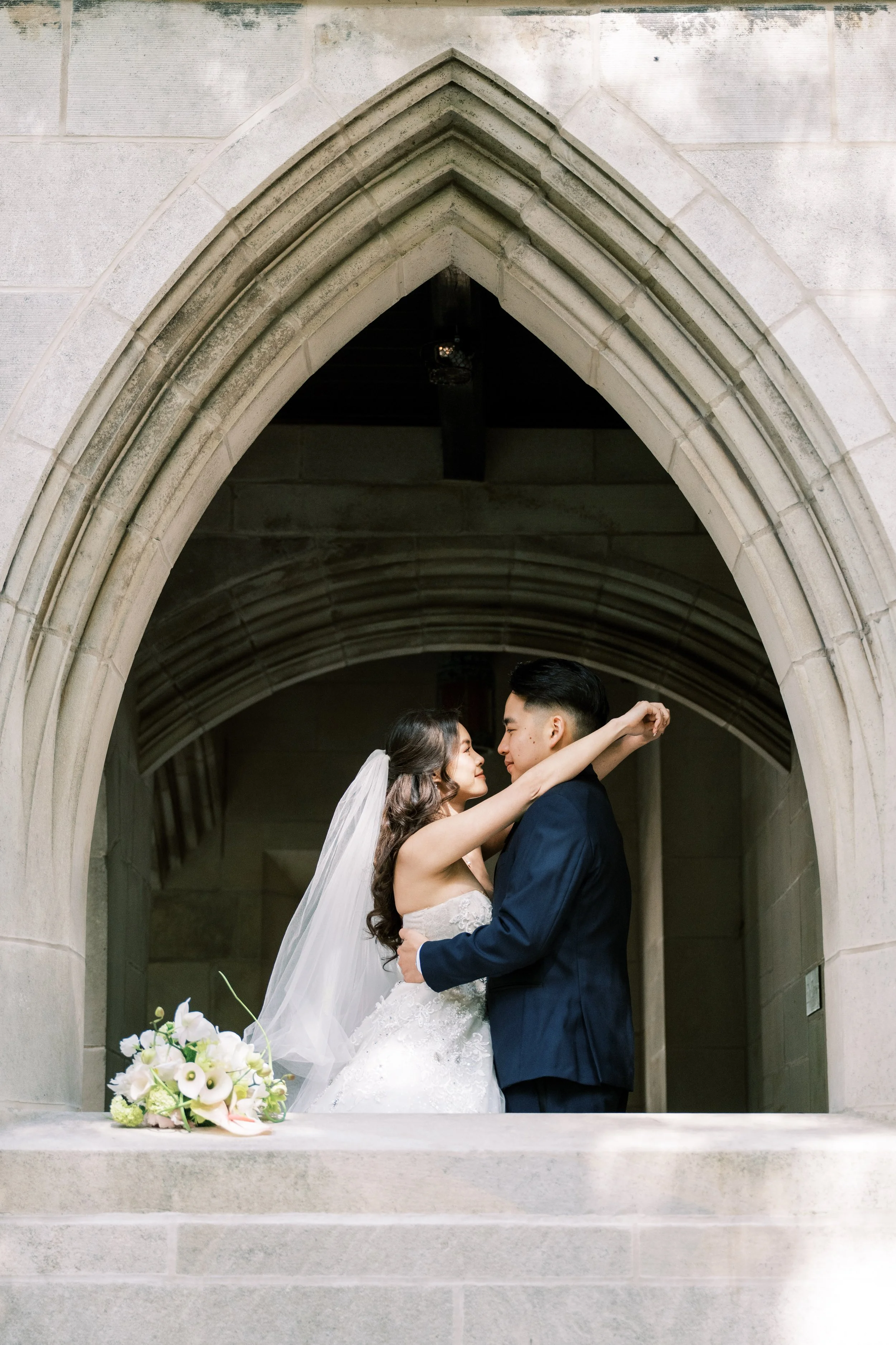 A bride and groom sharing a romantic moment under a stone archway. The bride is in a white wedding gown with a veil, and the groom is in a navy suit. A bouquet of white and green flowers is on the table in front of them.