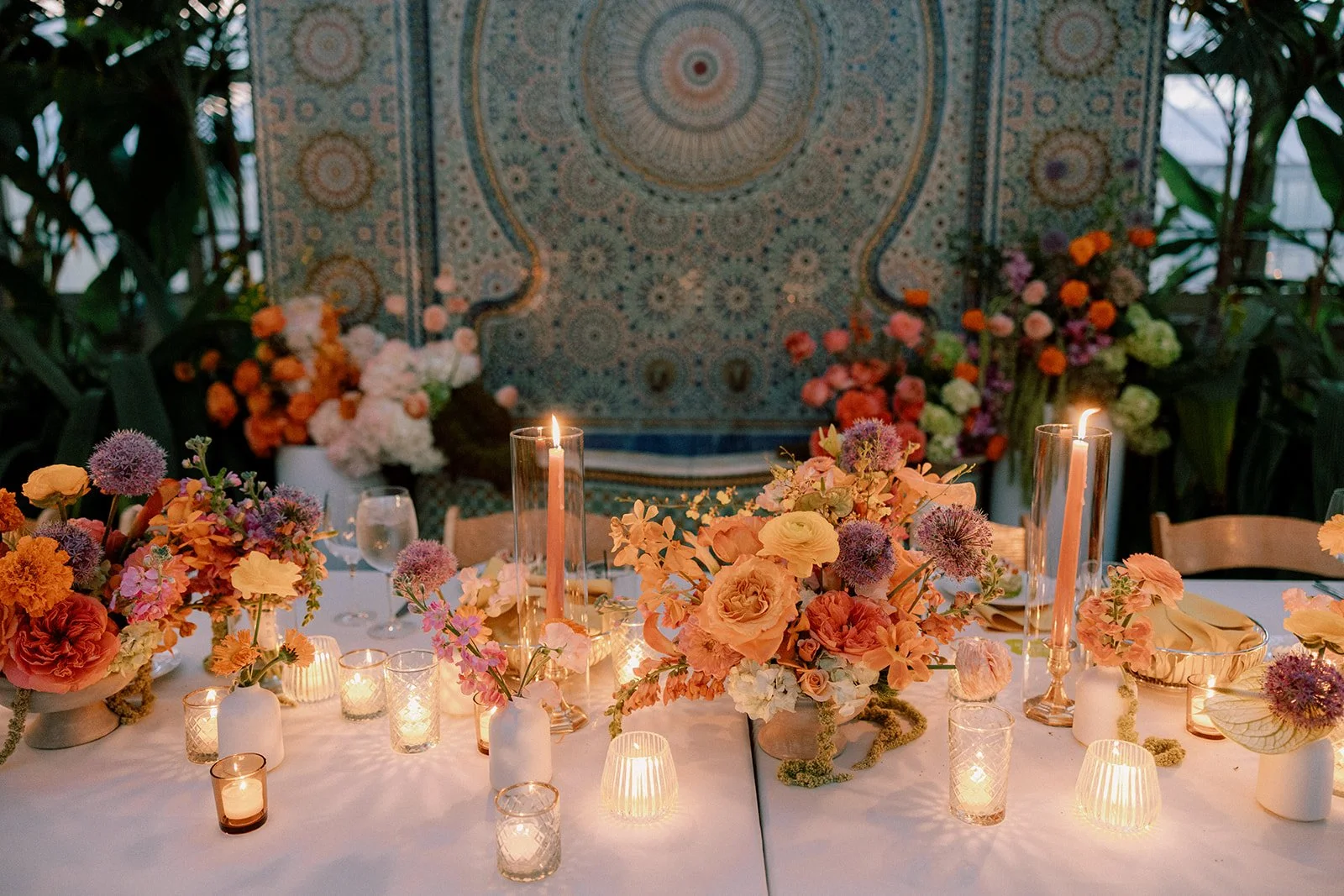 A beautifully decorated table with floral centerpieces, candles, and elegant table settings, set against a large patterned tapestry or wall hanging with lush greenery and flowers in the background.