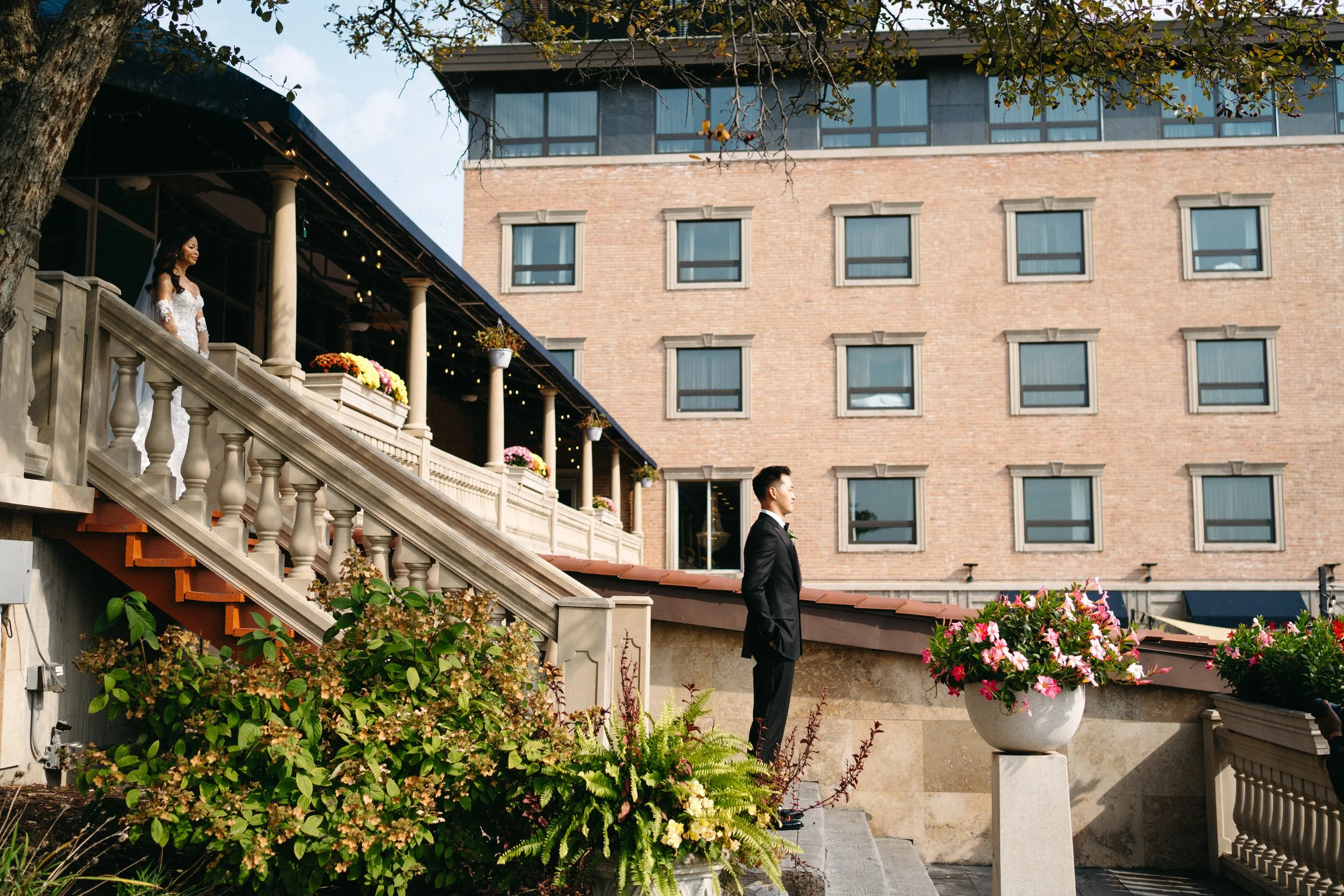 A bride in a white wedding dress standing on outdoor stairs decorated with flowers, with a groom in a suit standing nearby, in front of a large brick building with multiple windows.