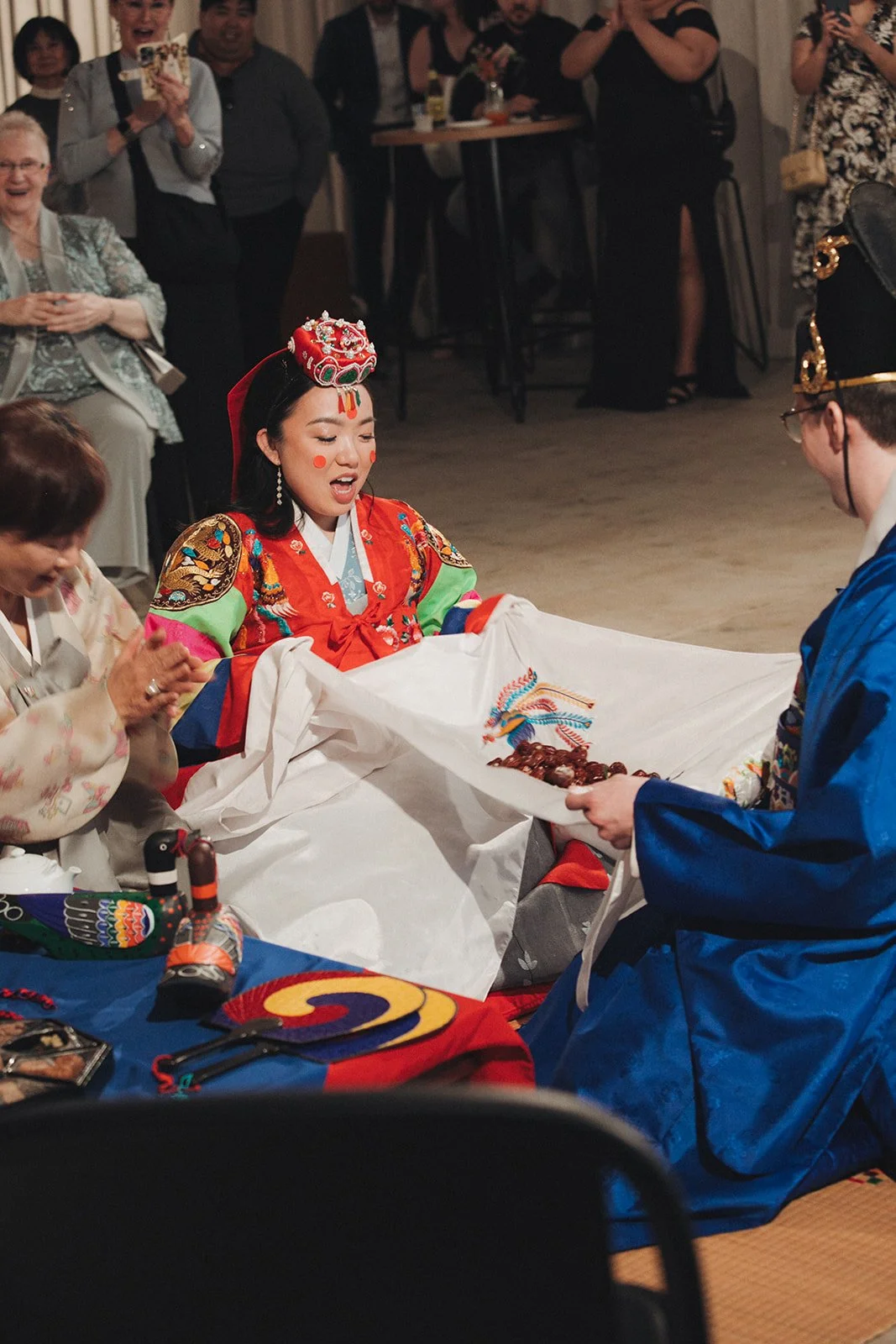 Korean bride in a colorful hanbok, kneeling during a traditional wedding ceremony, with guests watching and taking photos around her.
