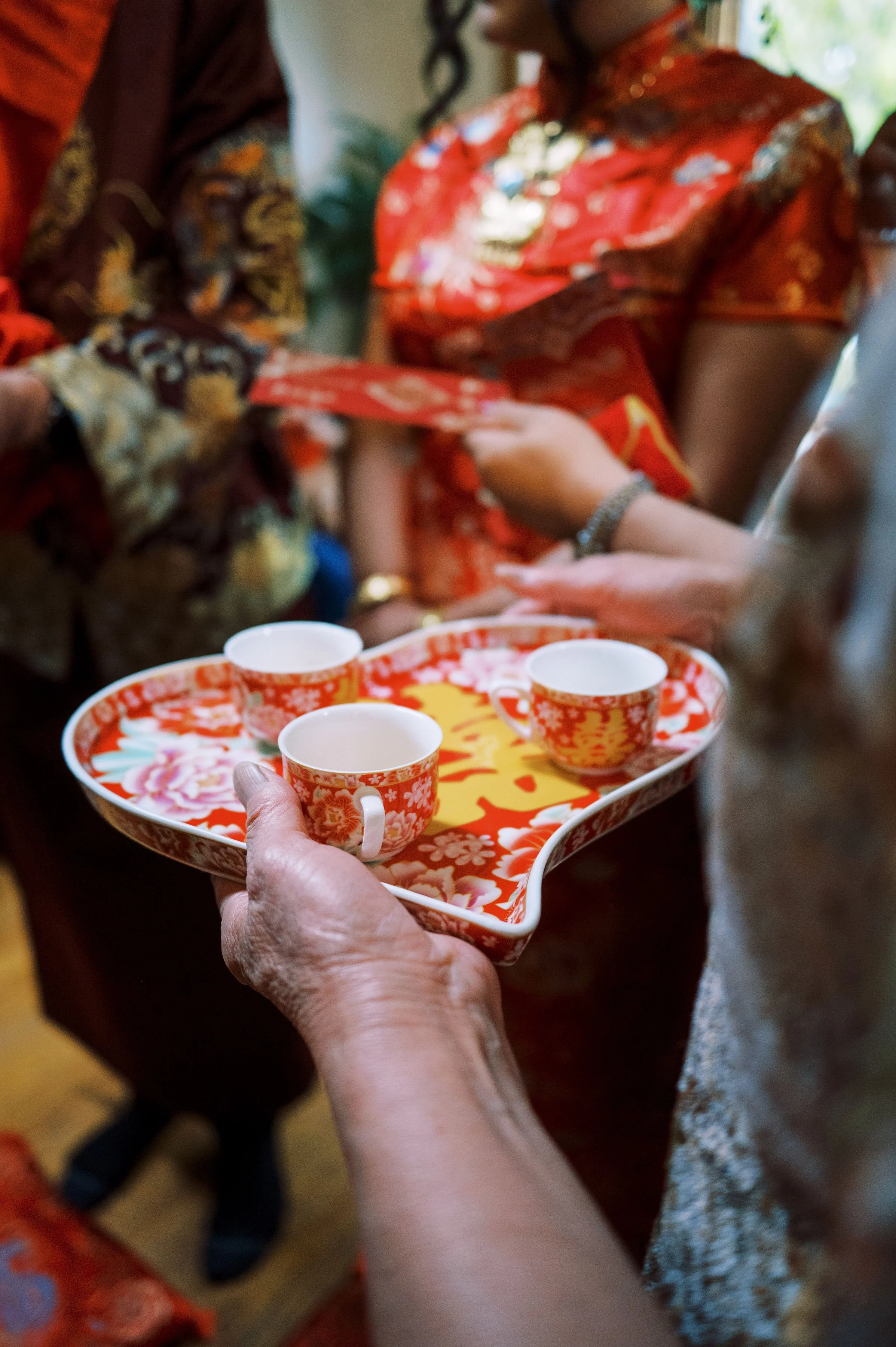 People participating in a traditional tea ceremony, with hands holding a tray of matching ceramic cups with floral patterns, set in a cultural setting with women wearing colorful dresses.