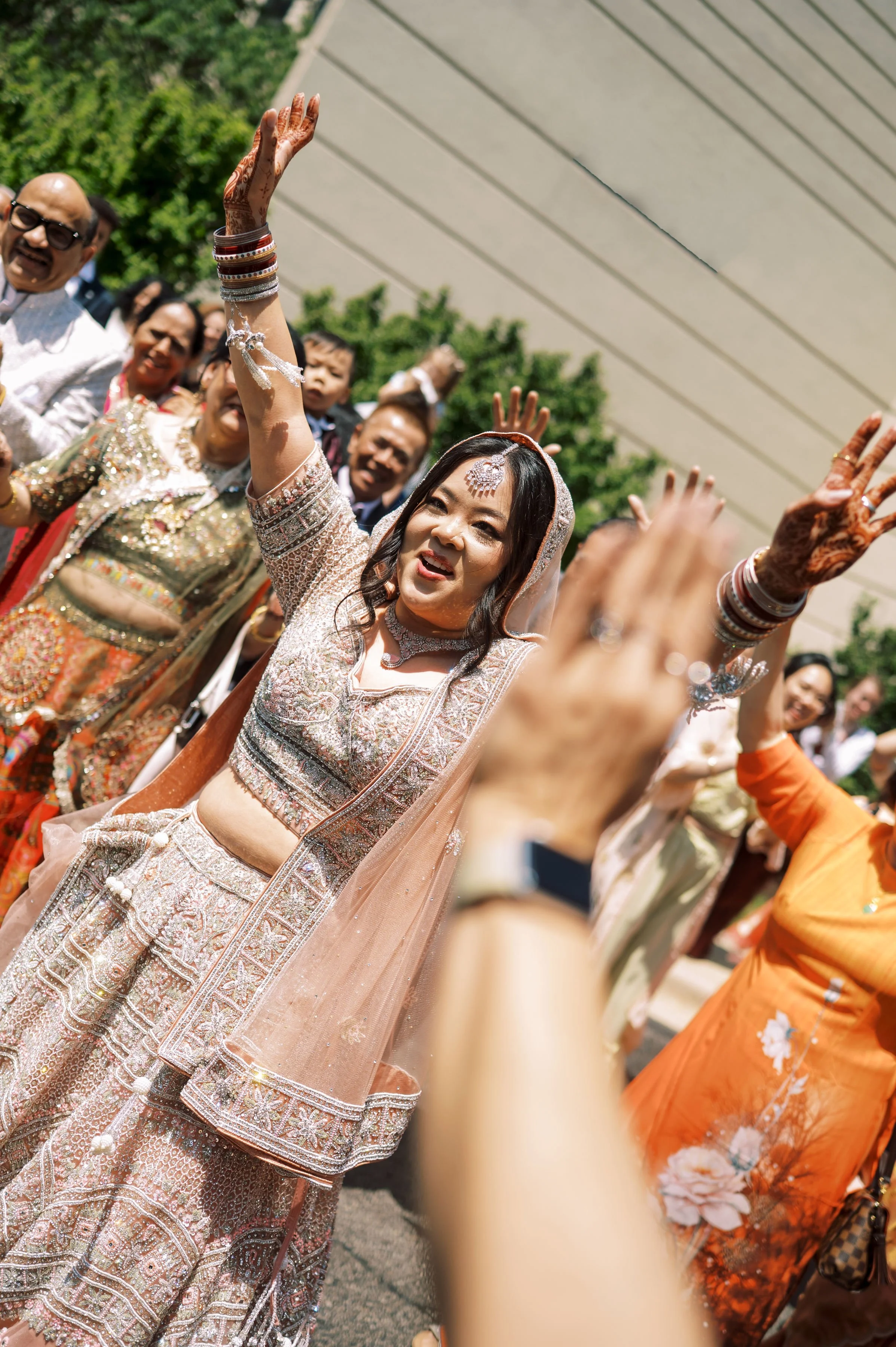 Group of people celebrating, with women in traditional Indian attire, including intricate embroidery and jewelry, raising their hands in the air, outdoors under a building overhang.