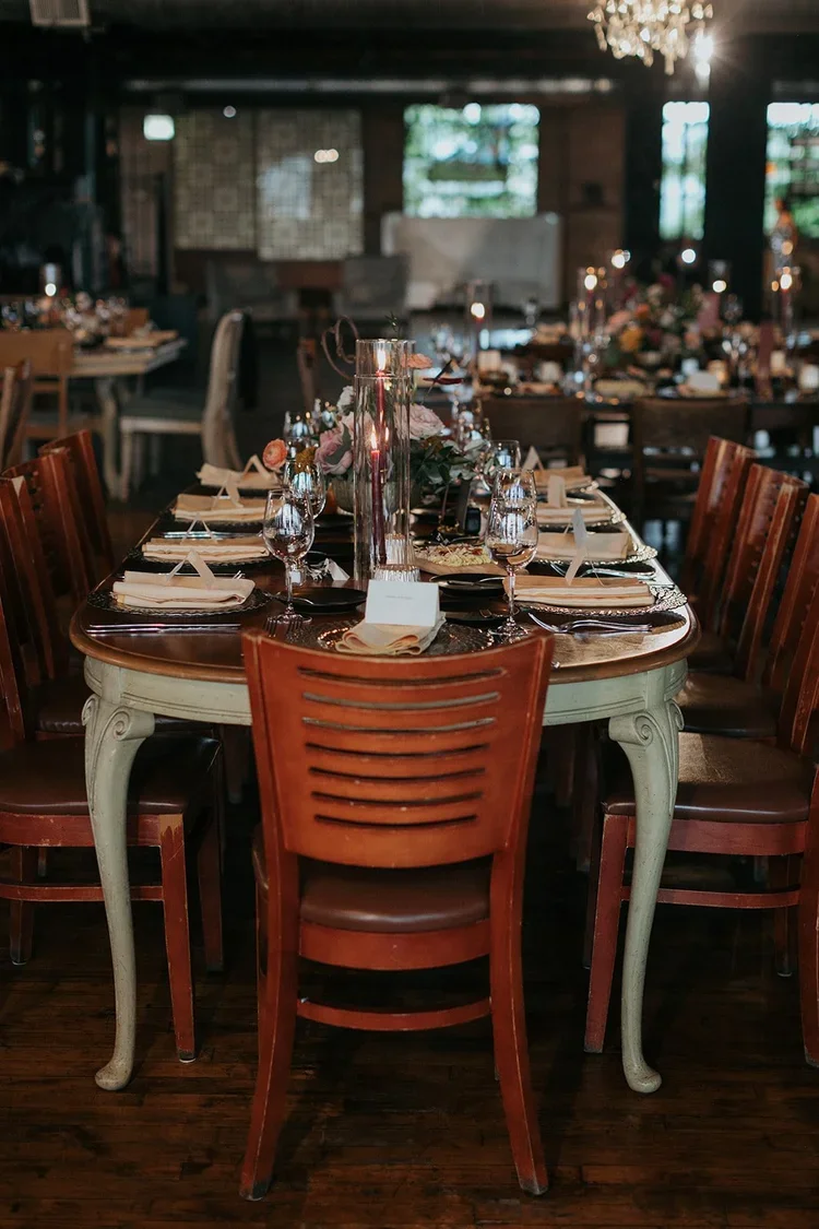 A decorated banquet table set for a formal event featuring glassware, plates, napkins, and floral centerpieces in a dimly lit restaurant or event space.
