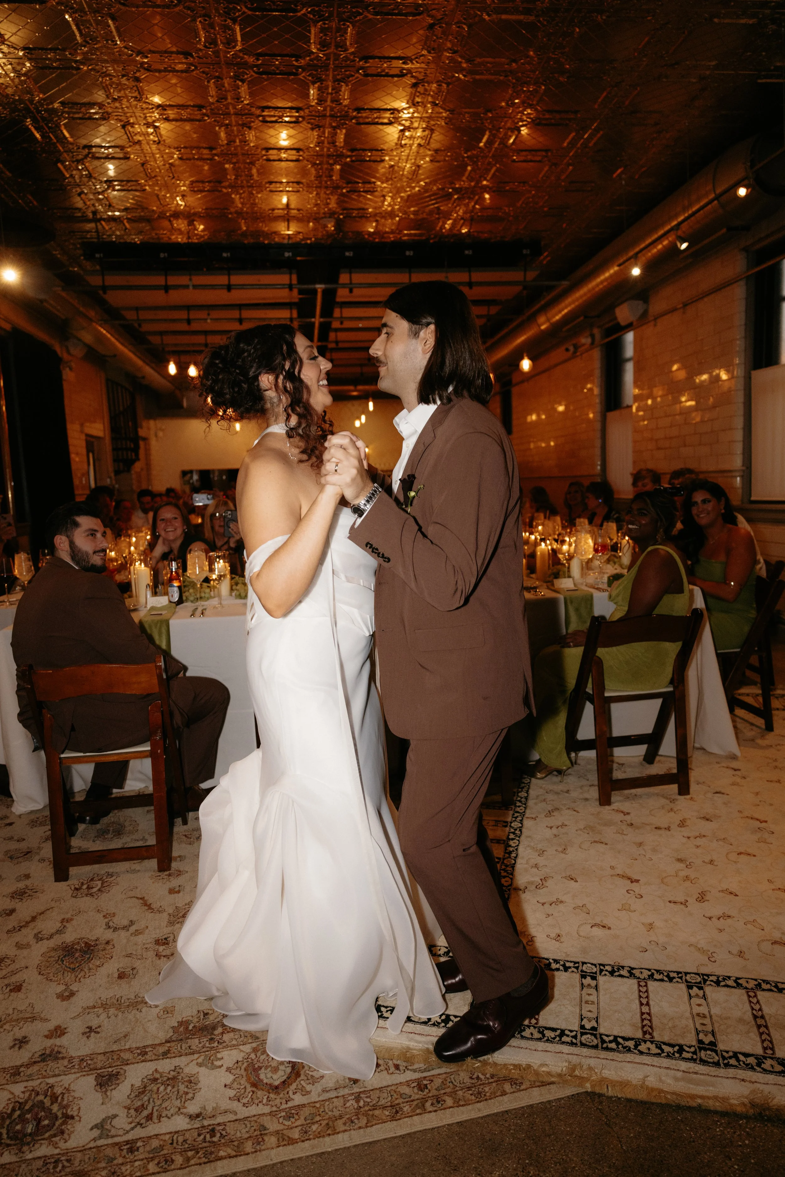 A couple dancing at a wedding reception. The bride is in a white gown, and the groom is wearing a brown suit. They are smiling and holding hands while dancing. Guests are seated at tables in the background, watching and smiling. The venue has warm li