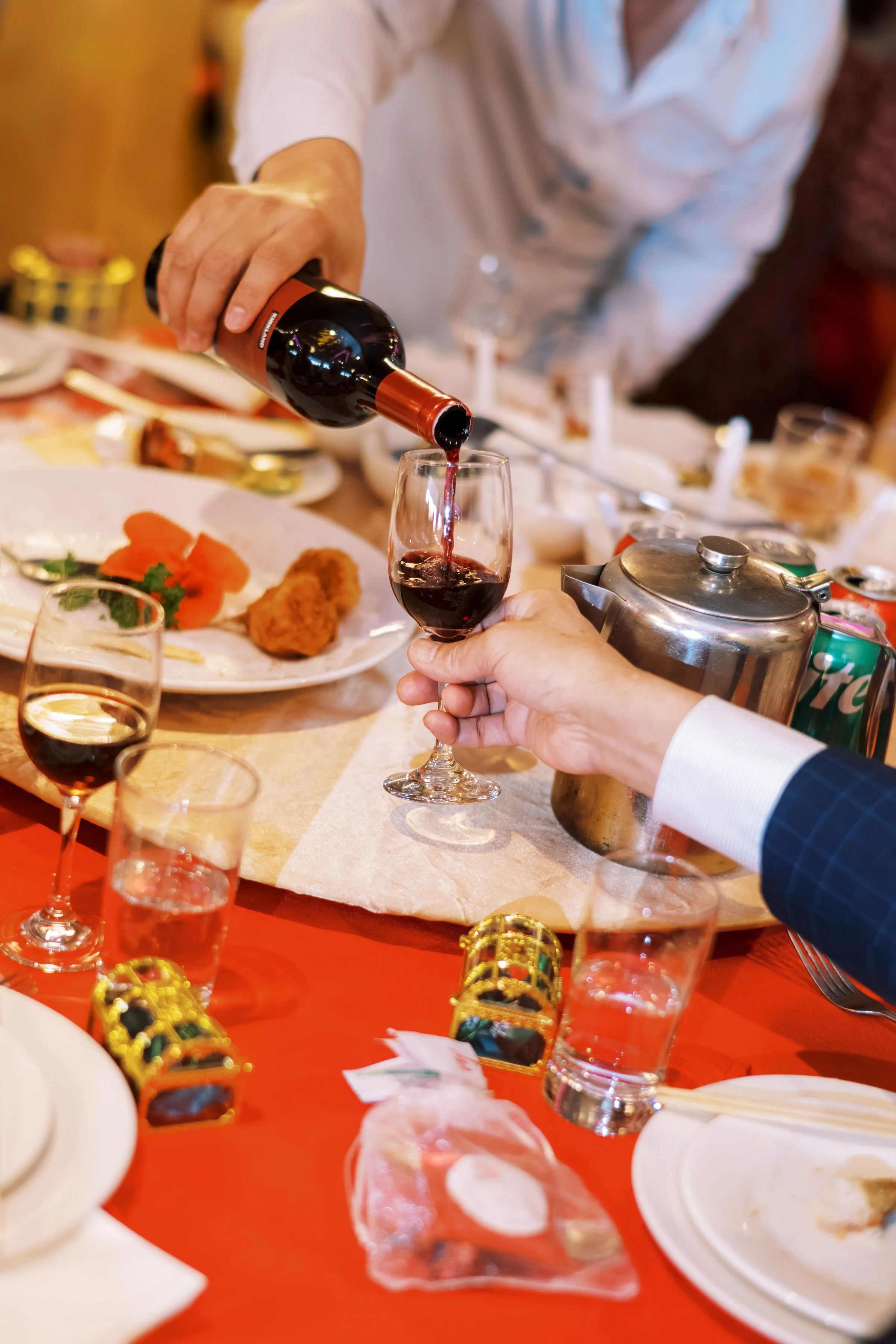 A person pouring red wine into a glass at a celebration or dinner event. The table is decorated with shiny decorations, food plates, glasses, and a teapot.