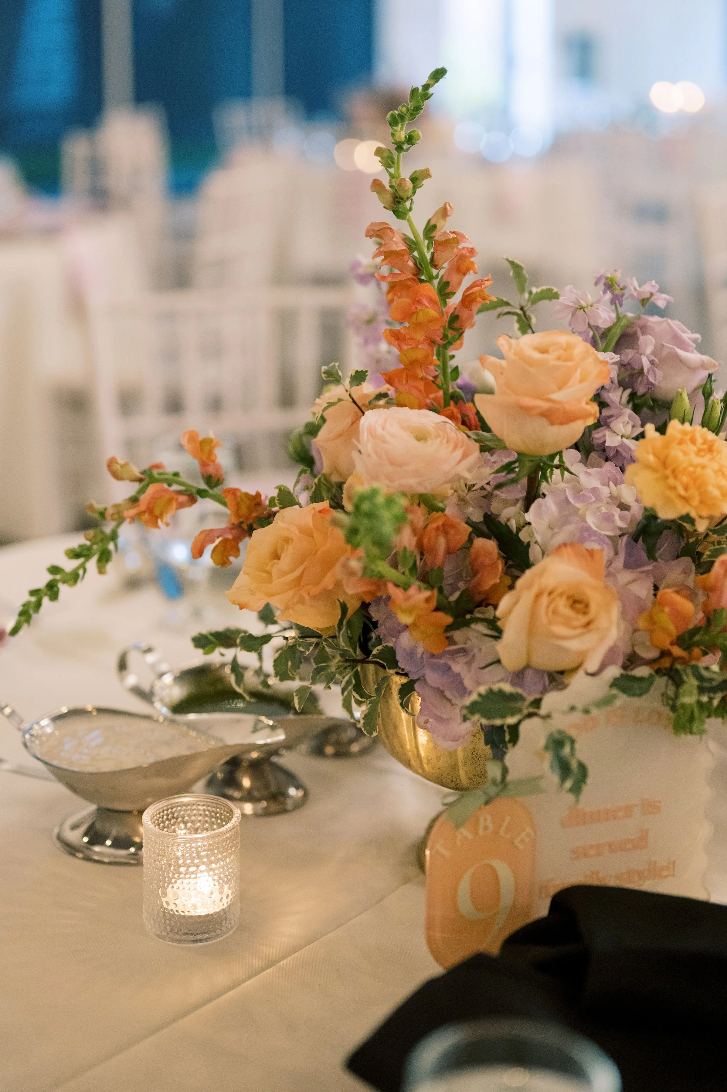 Close-up of a floral centerpiece with peach and lavender flowers on a table at an event, with a small lit candle and table setting visible in the foreground.