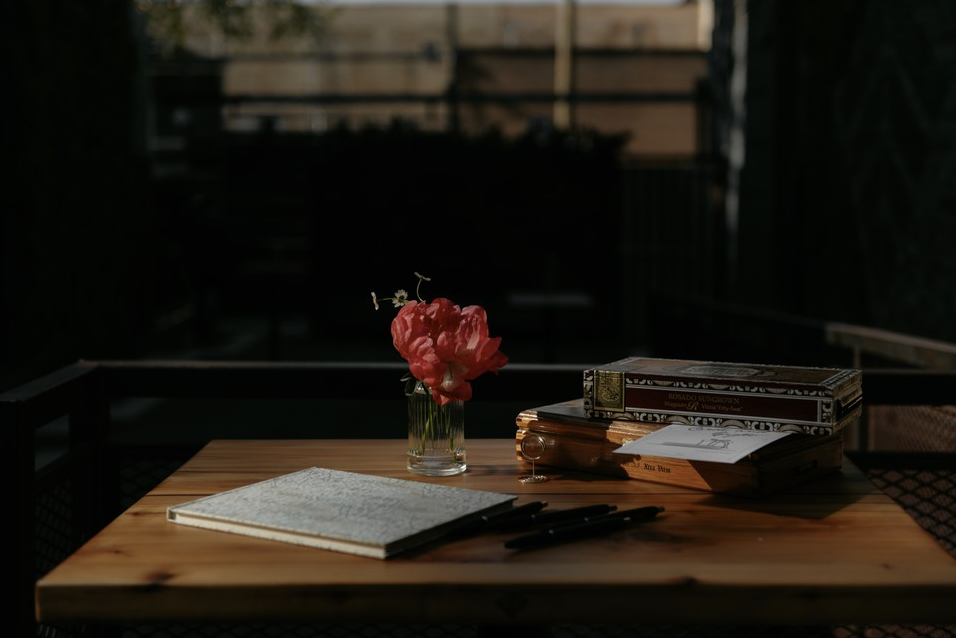 A wooden table with a small glass vase containing pink peonies, a closed notebook, three pens, and a stack of books, set outdoors with a dark, blurred background.