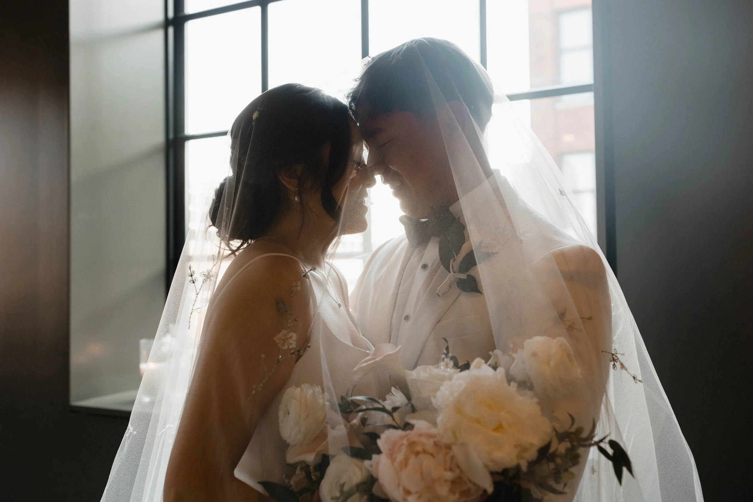 Two brides, wearing wedding dresses and veils, are about to kiss in front of a large window with natural light shining in.