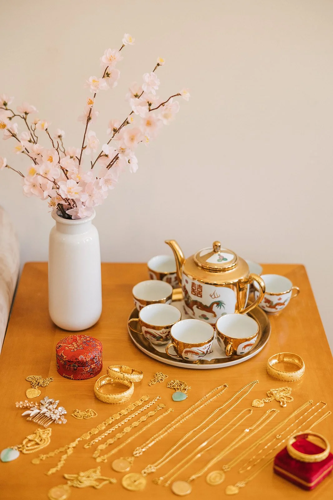 A wooden table displaying a white vase with pink cherry blossom branches, a golden-trimmed teapot with floral and dragon designs, six matching teacups with similar patterns on a tray, and various gold jewelry including bracelets, necklaces, and rings