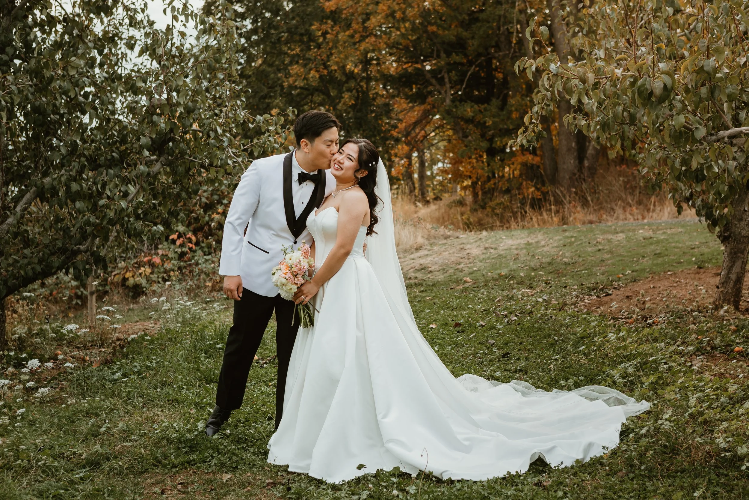 A newlywed couple standing outdoors in a wooded area with autumn leaves. The groom is giving the bride a kiss on the cheek. The bride is smiling, holding a bouquet. The bride wears a white wedding dress and veil, and the groom wears a white tuxedo ja