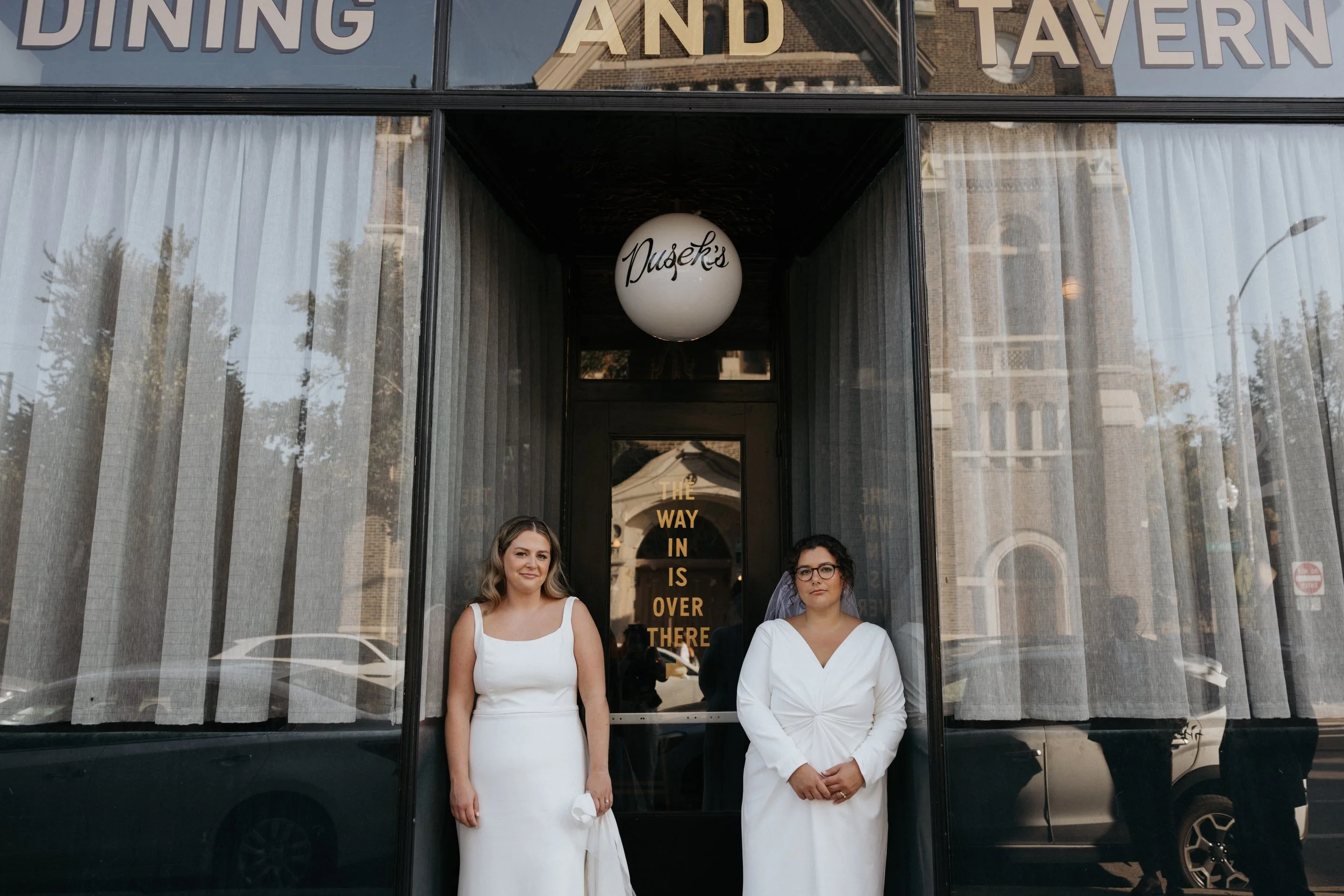 Two women stand in front of a glass window outside a restaurant or tavern. The woman on the left has light skin, long light brown hair, and is wearing a sleeveless white dress. The woman on the right has darker skin, curly dark hair, glasses, and is 