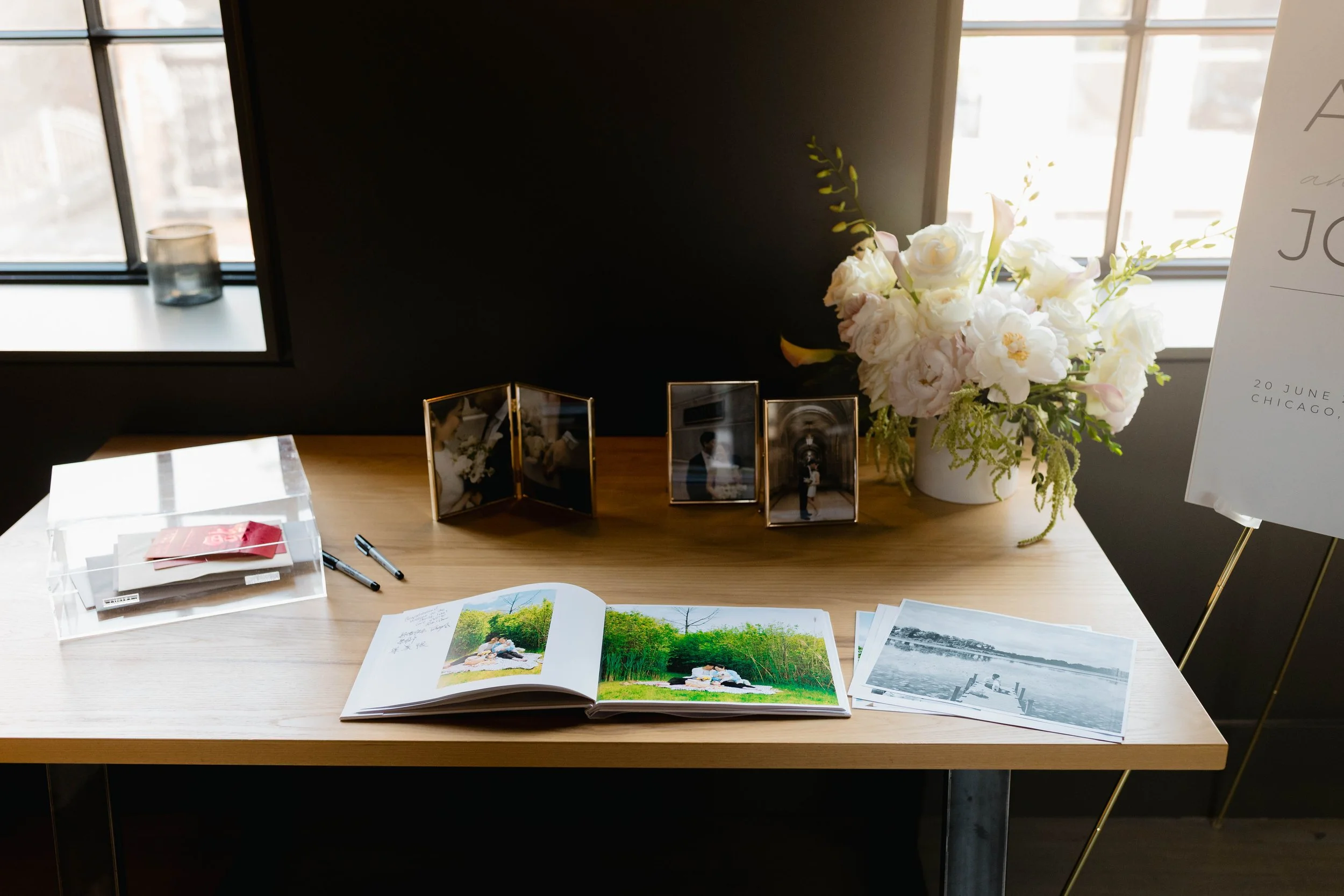 A table with an open photo album displaying a picture of two people outdoors, a clear plastic box with red and white paper inside, two black pens, and several black and white photographs. In the background, a bouquet of white flowers and greenery is 