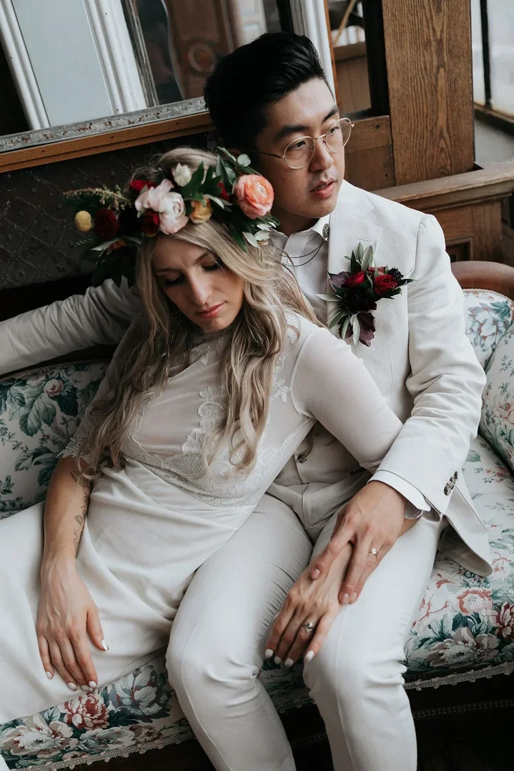 A couple in wedding attire sitting on a floral-patterned vintage sofa, with the bride wearing a floral crown and the groom wearing a boutonniere, in a cozy, rustic indoor setting.