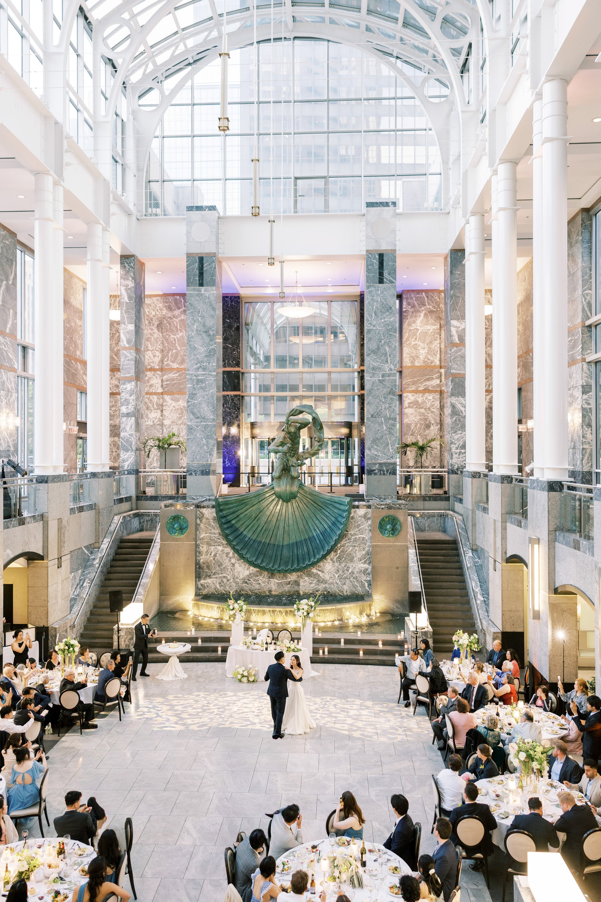 Wedding reception in a grand, modern atrium with high glass ceilings, a water fountain with a bronze statue, and elegantly dressed guests seated at round tables, with a couple dancing in the center.