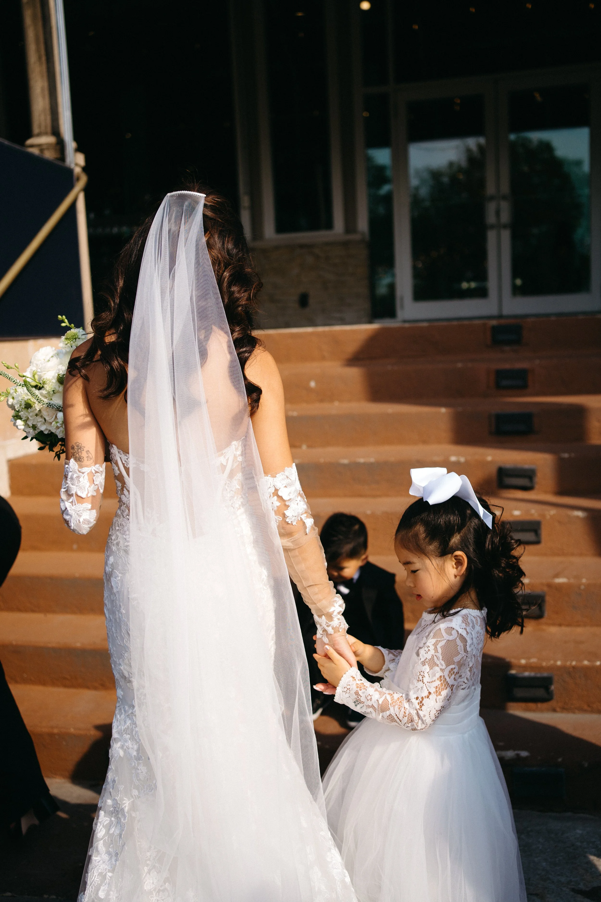 A bride and a young girl, possibly a flower girl, holding hands during a wedding ceremony. The bride is dressed in a white bridal gown with lace sleeves and a veil, while the girl wears a white lace dress with a large bow in her hair. They are standi