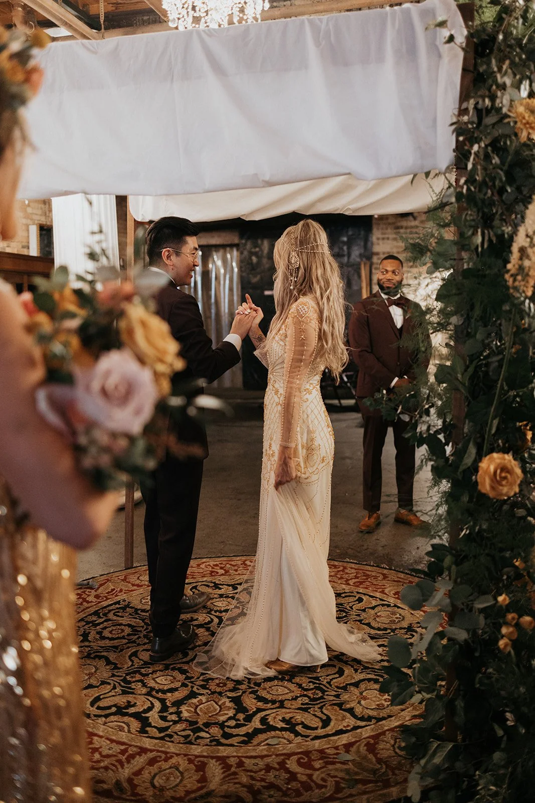 A couple exchanging vows at a wedding ceremony, with a man in a tuxedo standing nearby, decorated with floral arrangements and a patterned rug.