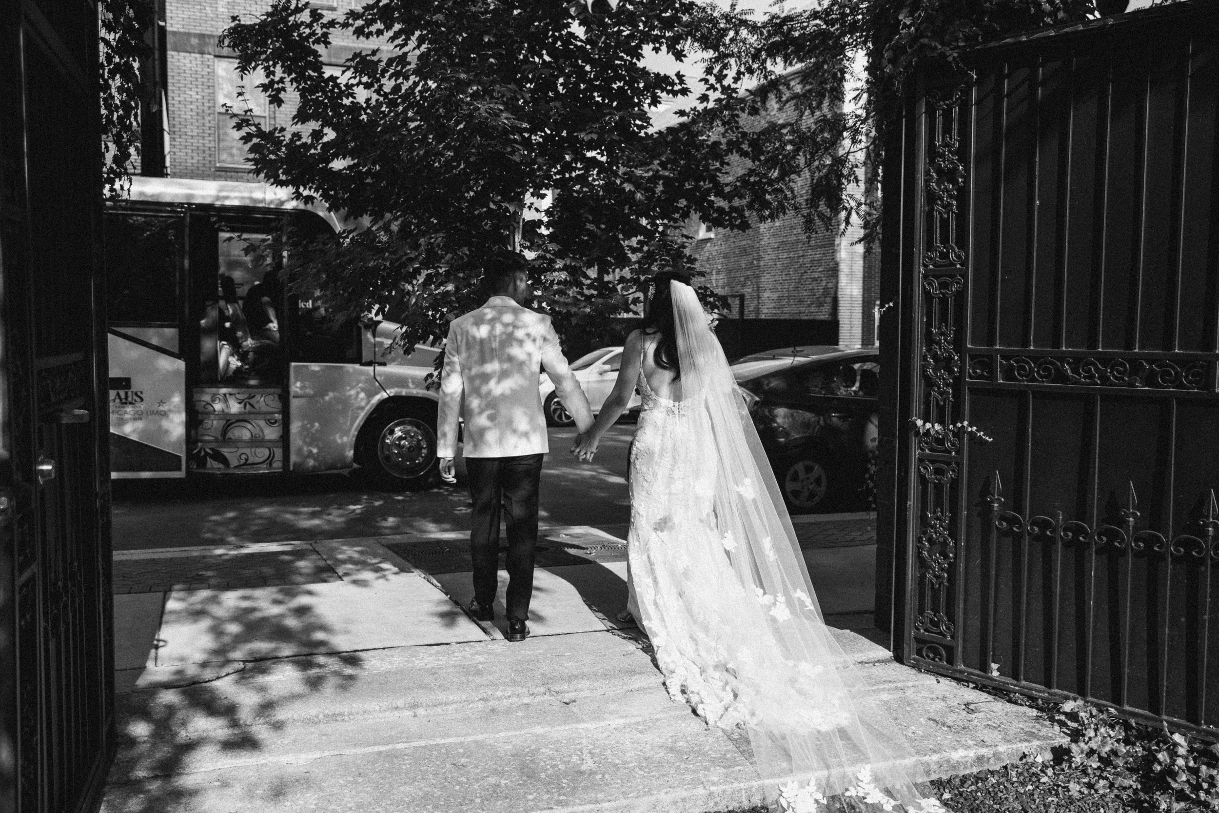 A bride and groom holding hands walking through an open wrought-iron gate onto a city street, with a tree and parked cars visible in the background, in black and white.