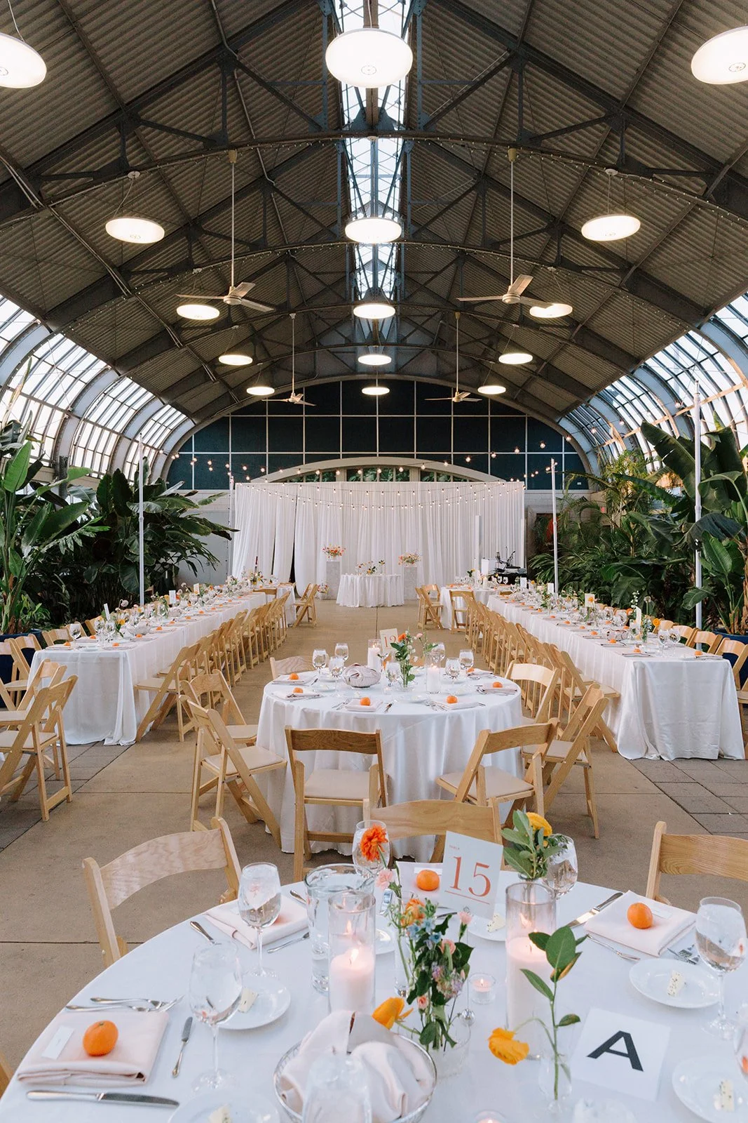 Elegant indoor wedding reception setup with long banquet tables and round tables, decorated with white tablecloths, flowers, candles, and orange accents, under a curved glass ceiling with hanging lights and ceiling fans.